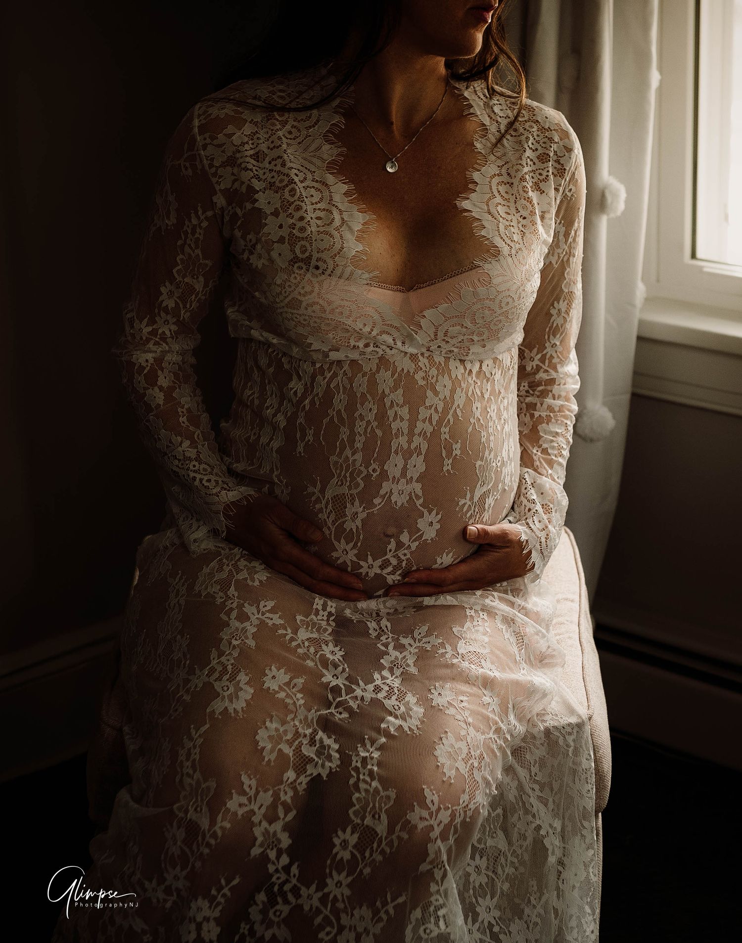A pregnant woman cradles her baby bump wearing an elegant white lace maternity gown near a sunlit window.