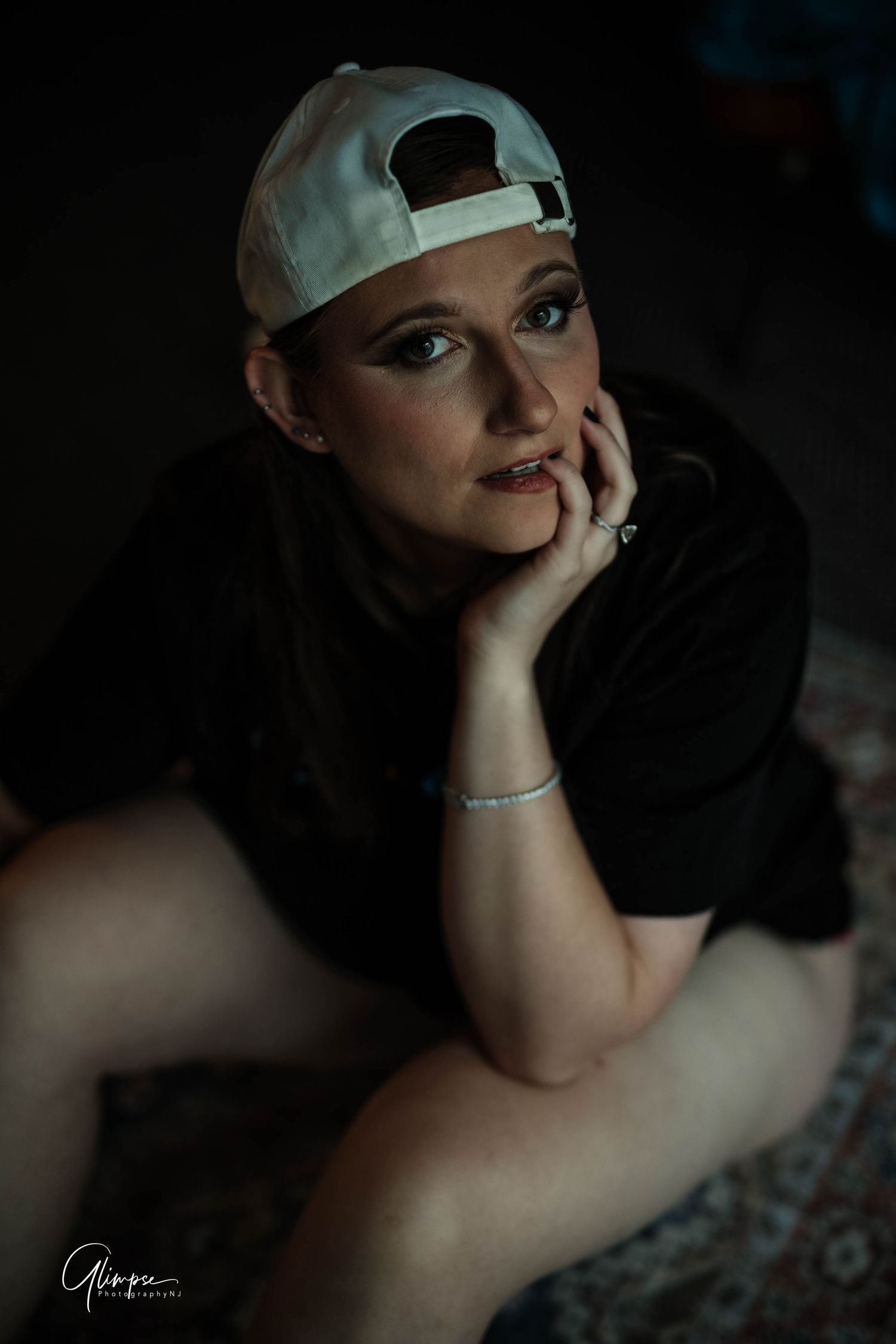 Young woman in a black t-shirt and backwards cap sitting on the floor in moody lighting.