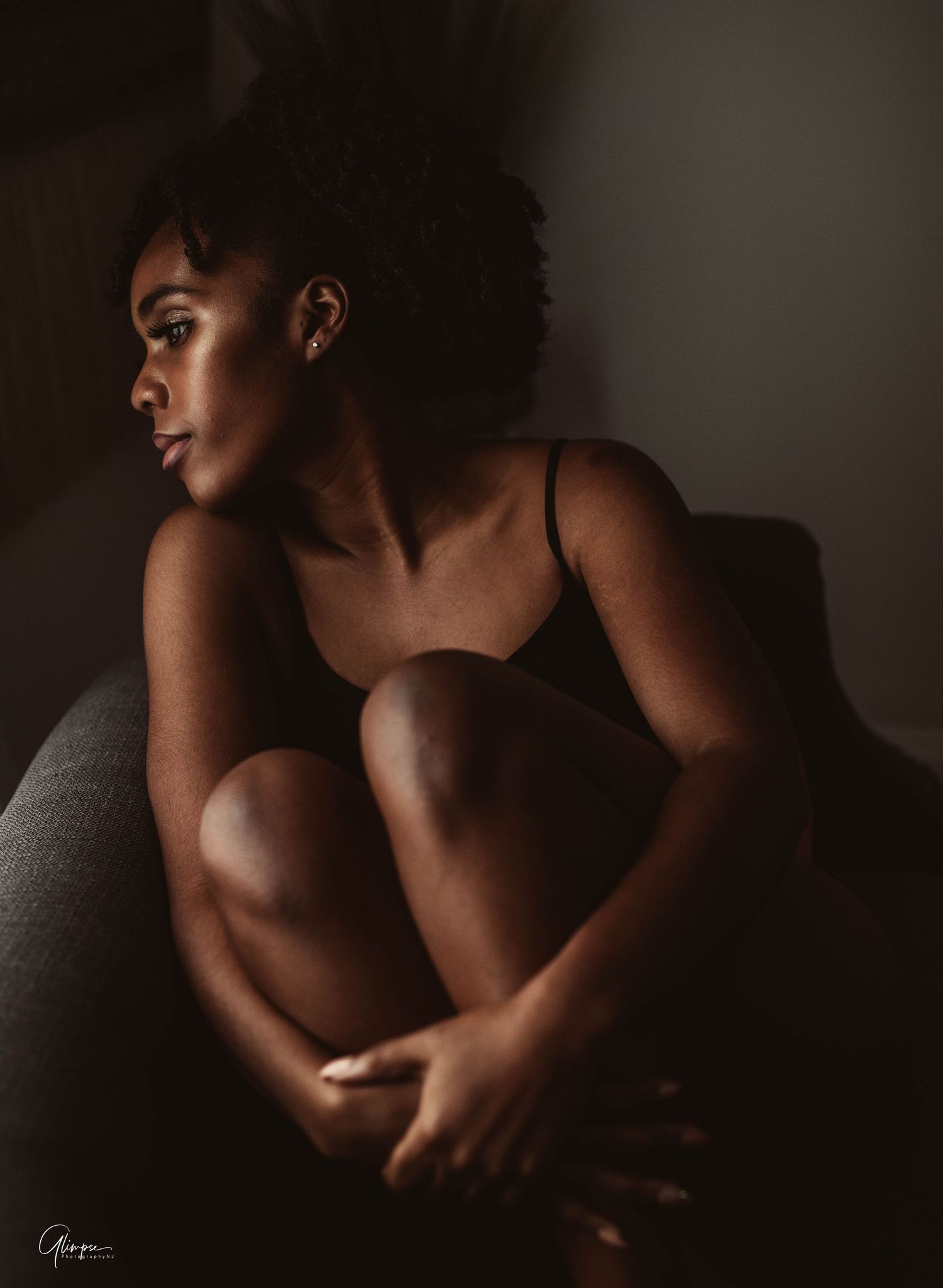 A young Black woman with natural hair sits curled up on a couch in moody dramatic lighting wearing a black bodysuit.