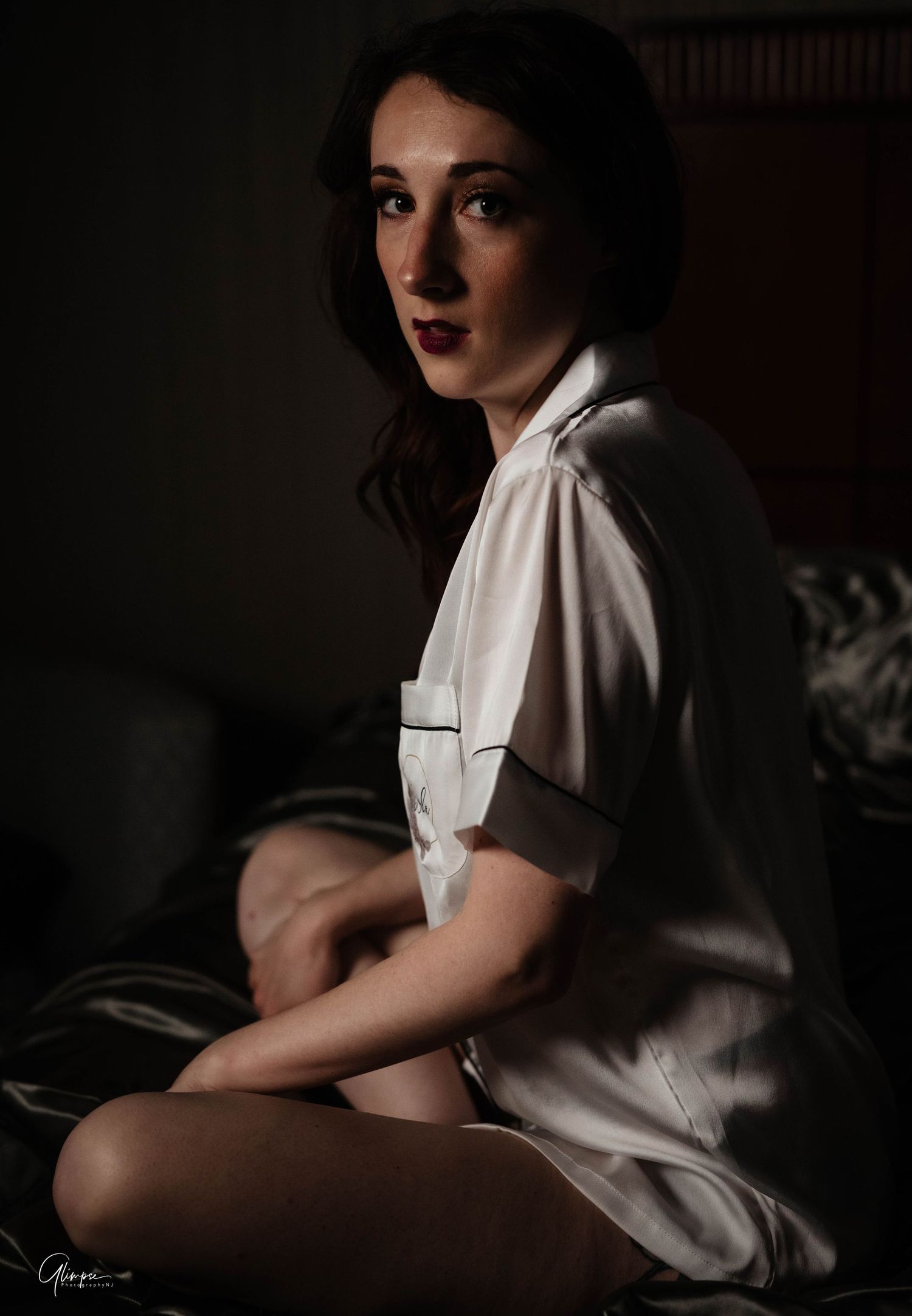 Young dark-haired woman in white satin shirt poses in moody low-light boudoir portrait.