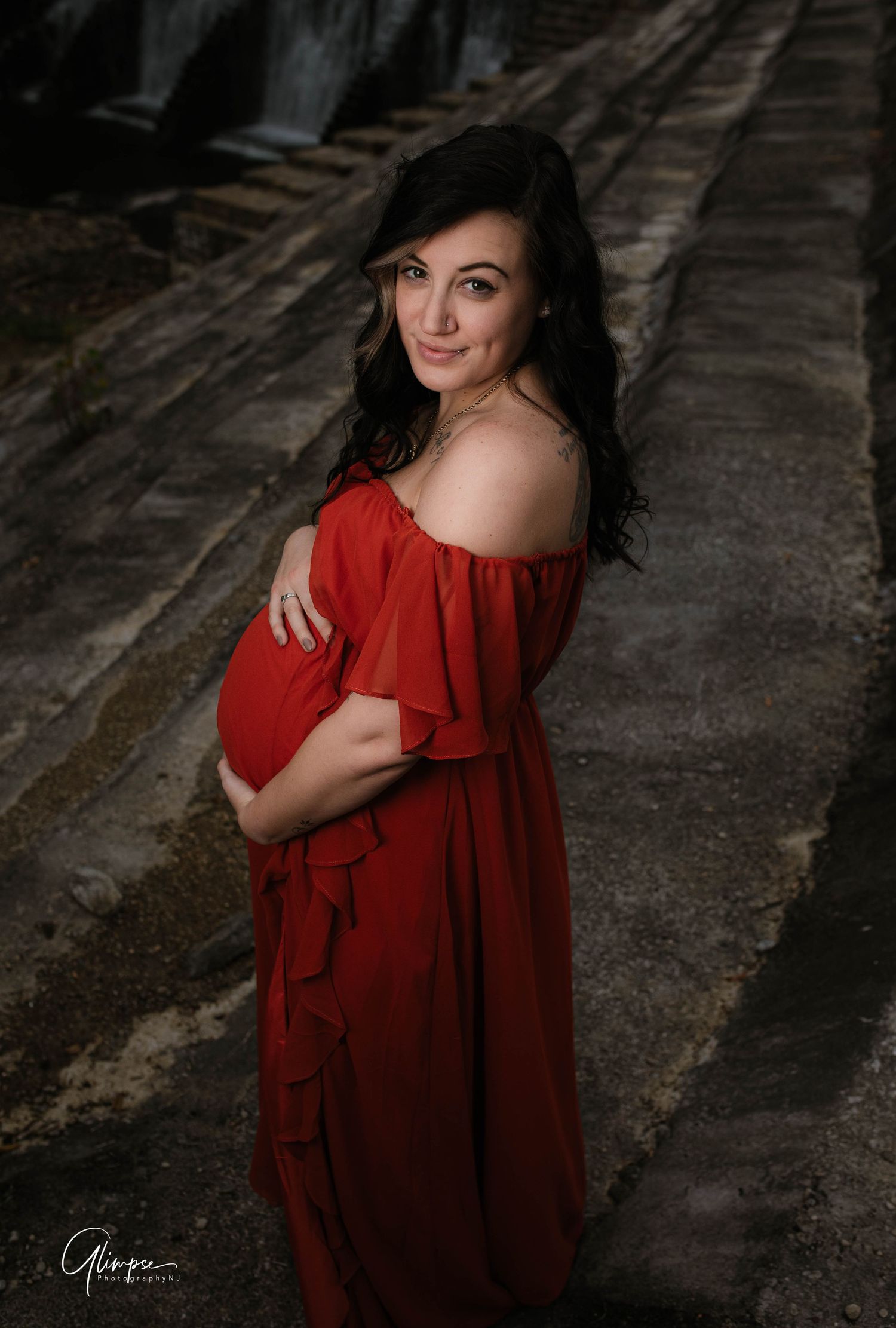 Beautiful pregnant woman in red off-shoulder gown posing gracefully during an outdoor maternity photography session.