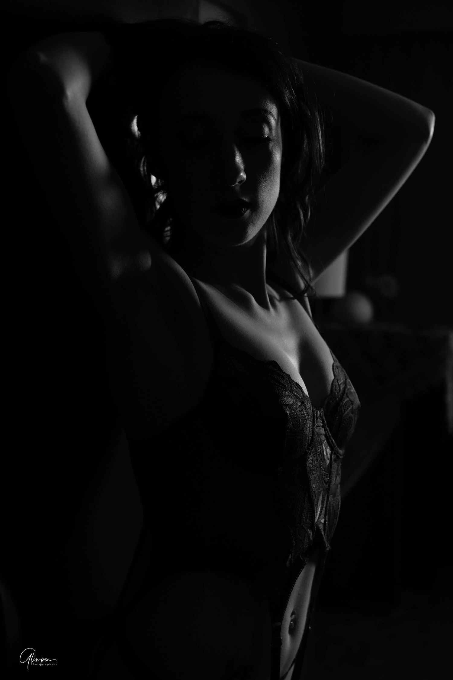 Black and white silhouette of woman in lace lingerie with hands raised in dark setting.