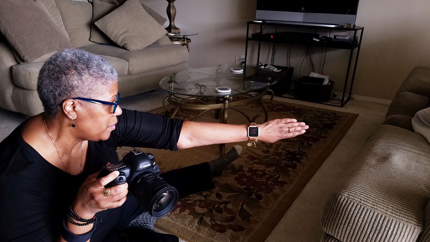 A woman with short gray hair holds a camera while extending her arm in a cozy living room setting.