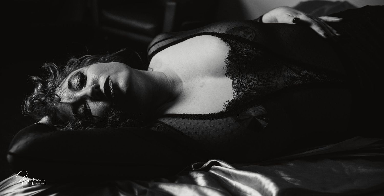 Black and white boudoir photo of a woman lying on satin sheets in dark lace lingerie.