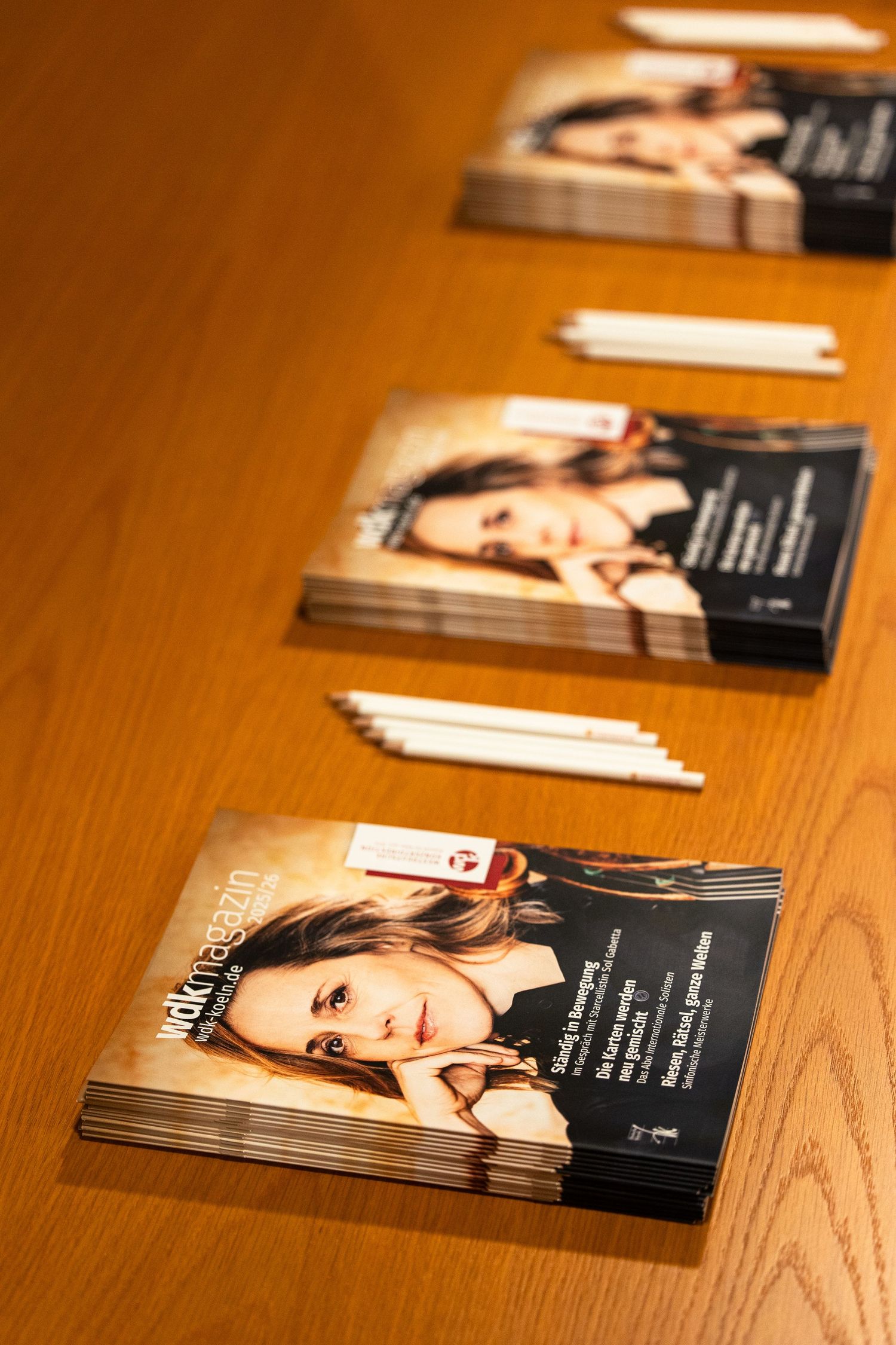 Stacks of wak magazin publications featuring a woman on the cover arranged in a row on a wooden table with pencils.