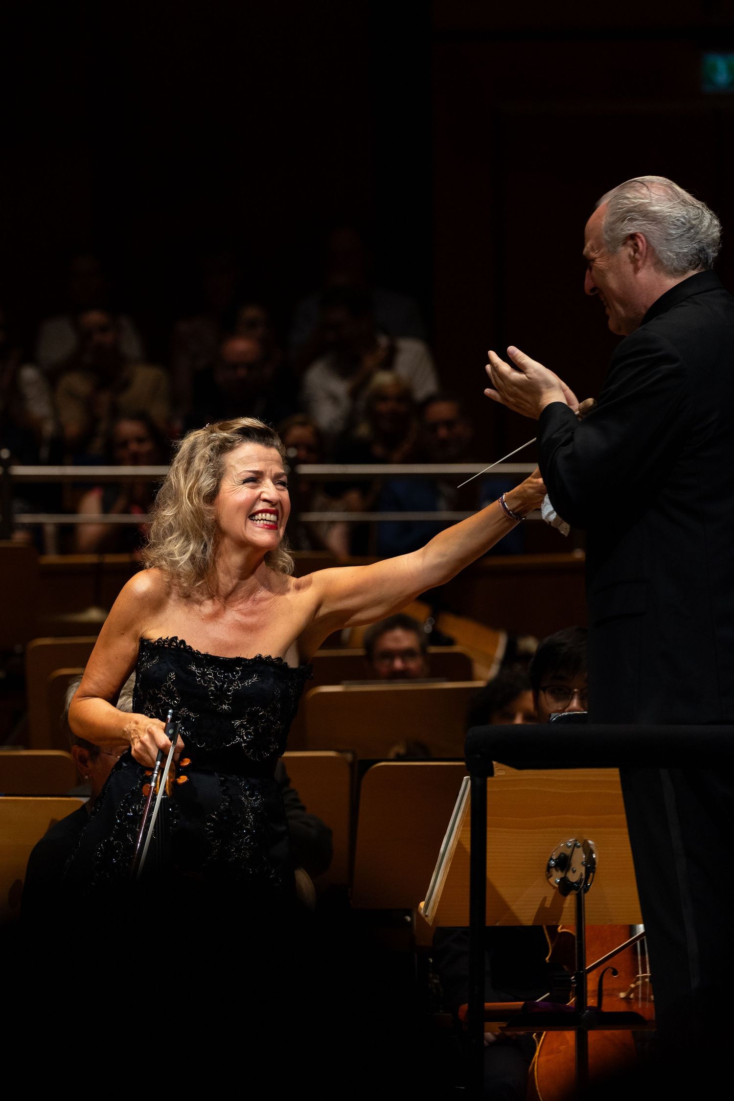 Female violinist in black gown reaches toward conductor with baton during orchestral performance on concert hall stage.