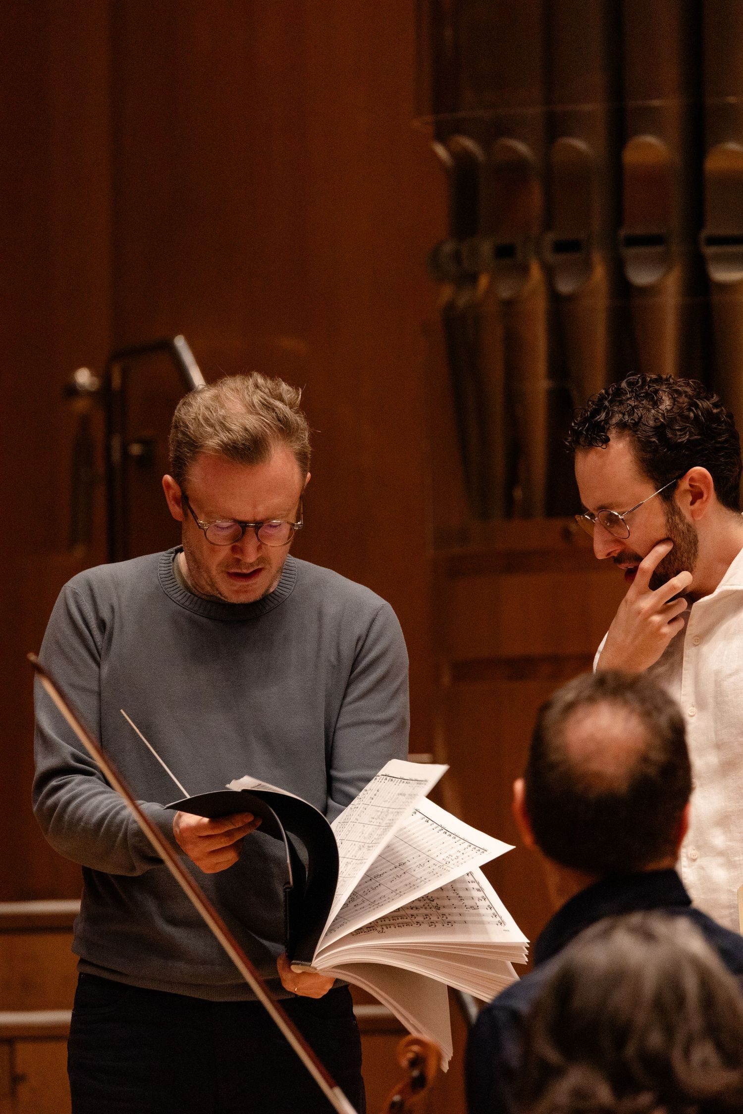 Two musicians reviewing sheet music together in a concert hall with organ pipes visible in the background.