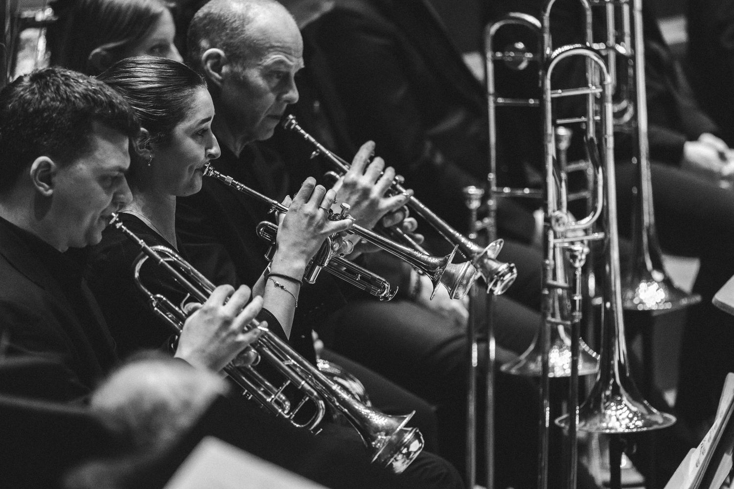 Black and white photo of orchestra musicians playing trumpets and trombones during a live performance.