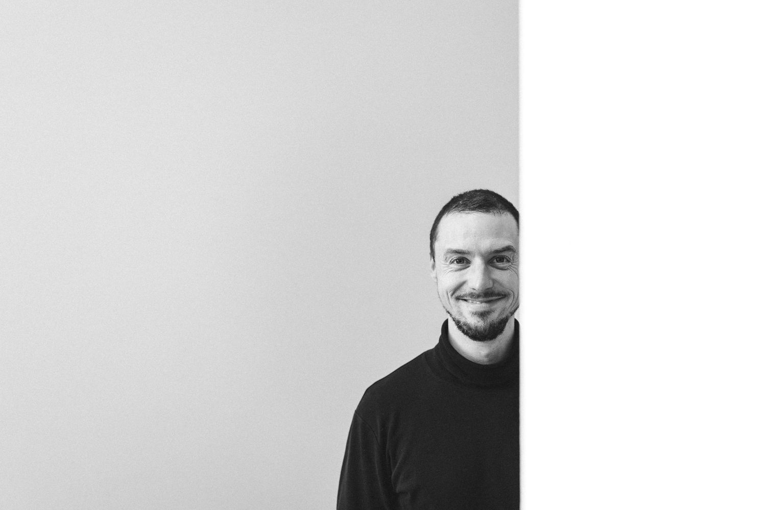 Smiling man with beard wearing black turtleneck, peeking from behind white wall in black and white photo.