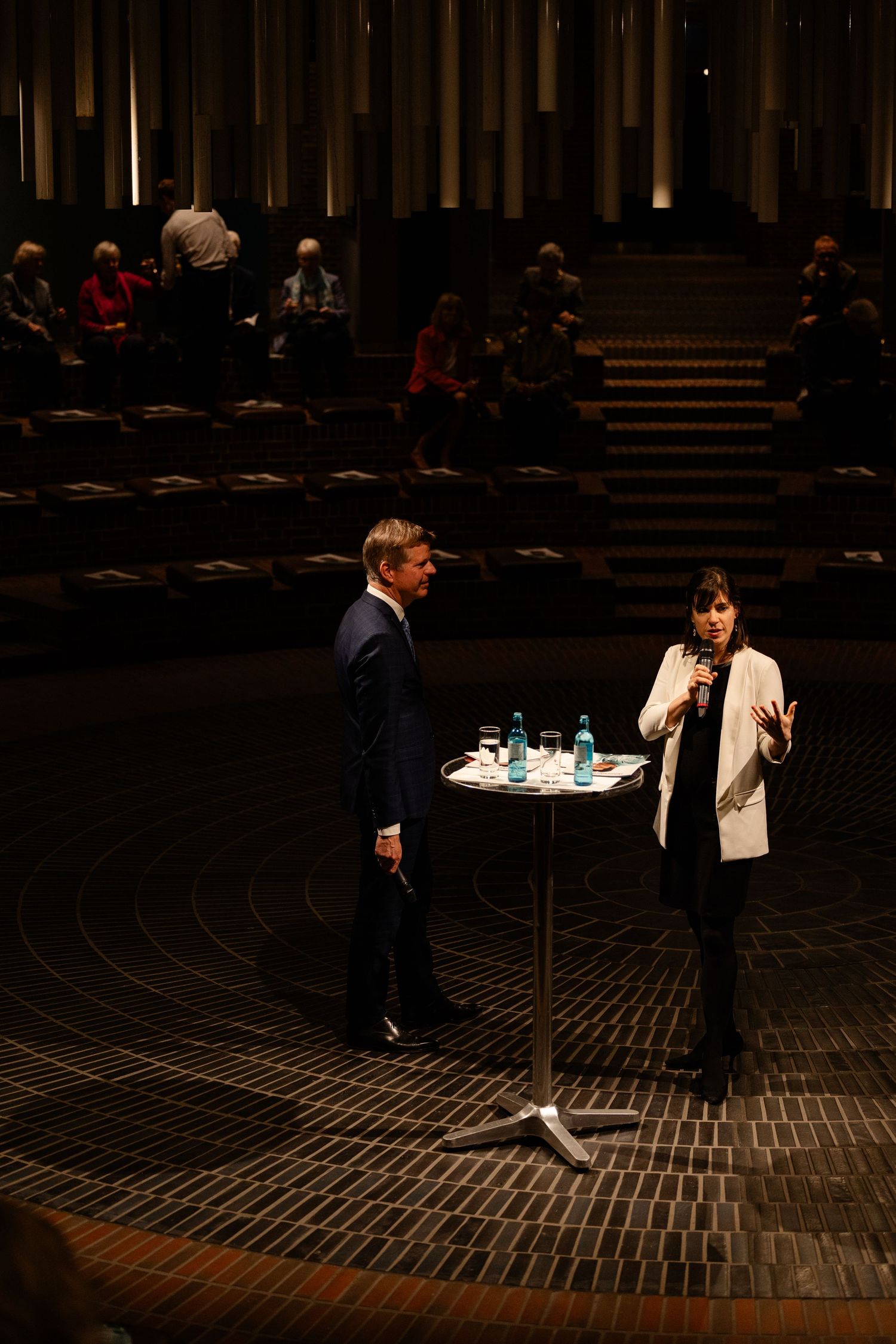 Two speakers stand at a high table in a dimly lit auditorium during a formal panel discussion event.