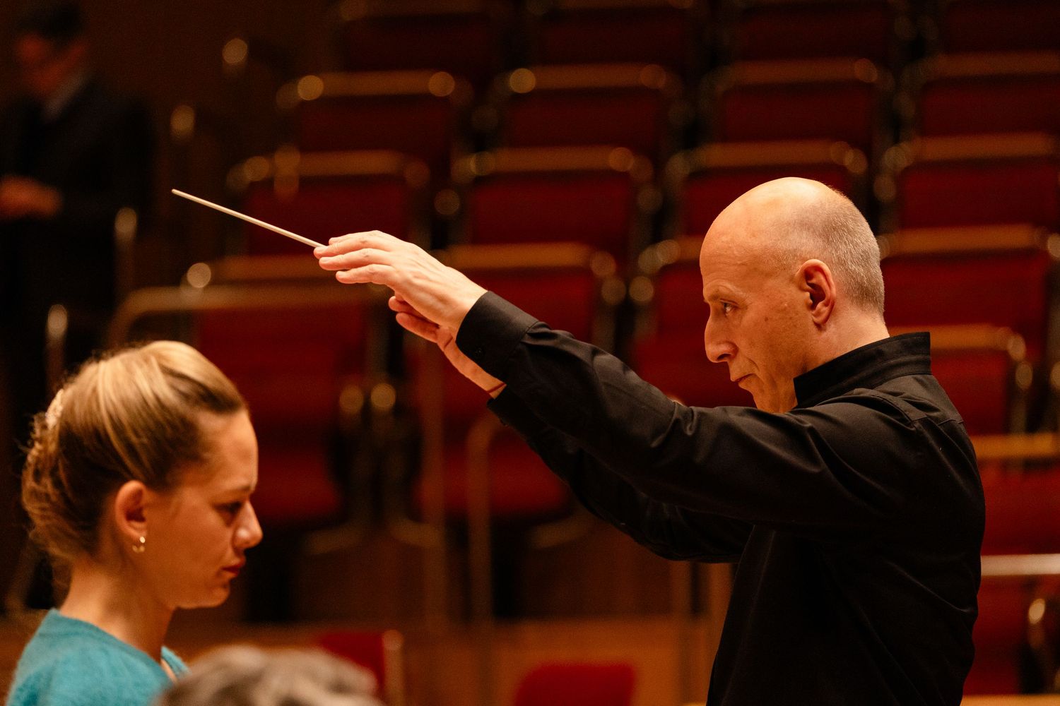 A bald conductor raises a baton while leading a young female musician in a concert hall.
