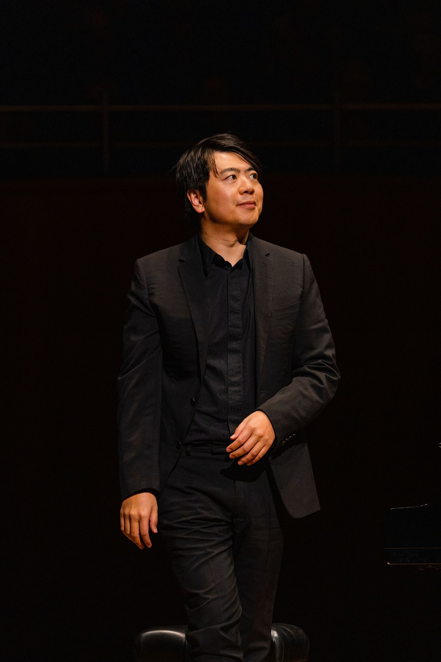 Man in black suit and shirt stands confidently on a dimly lit stage, gazing thoughtfully to the side.