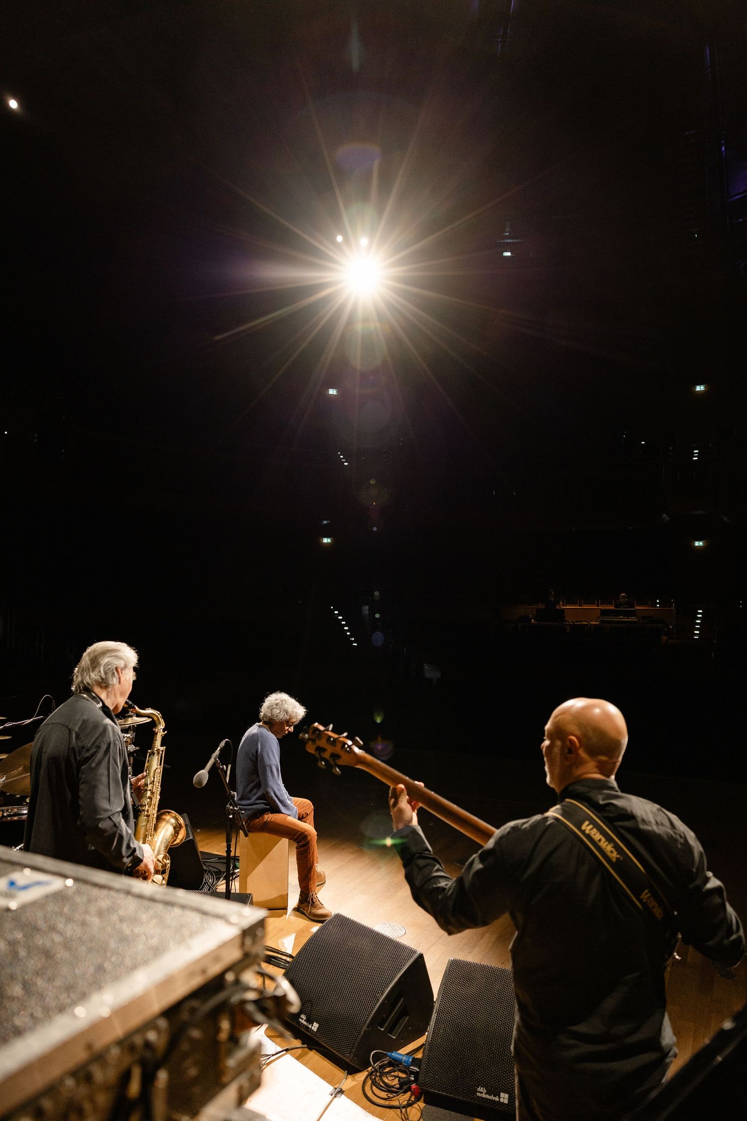 Three musicians perform on a dimly lit stage, with a saxophonist, bassist, and guitarist under a bright spotlight.