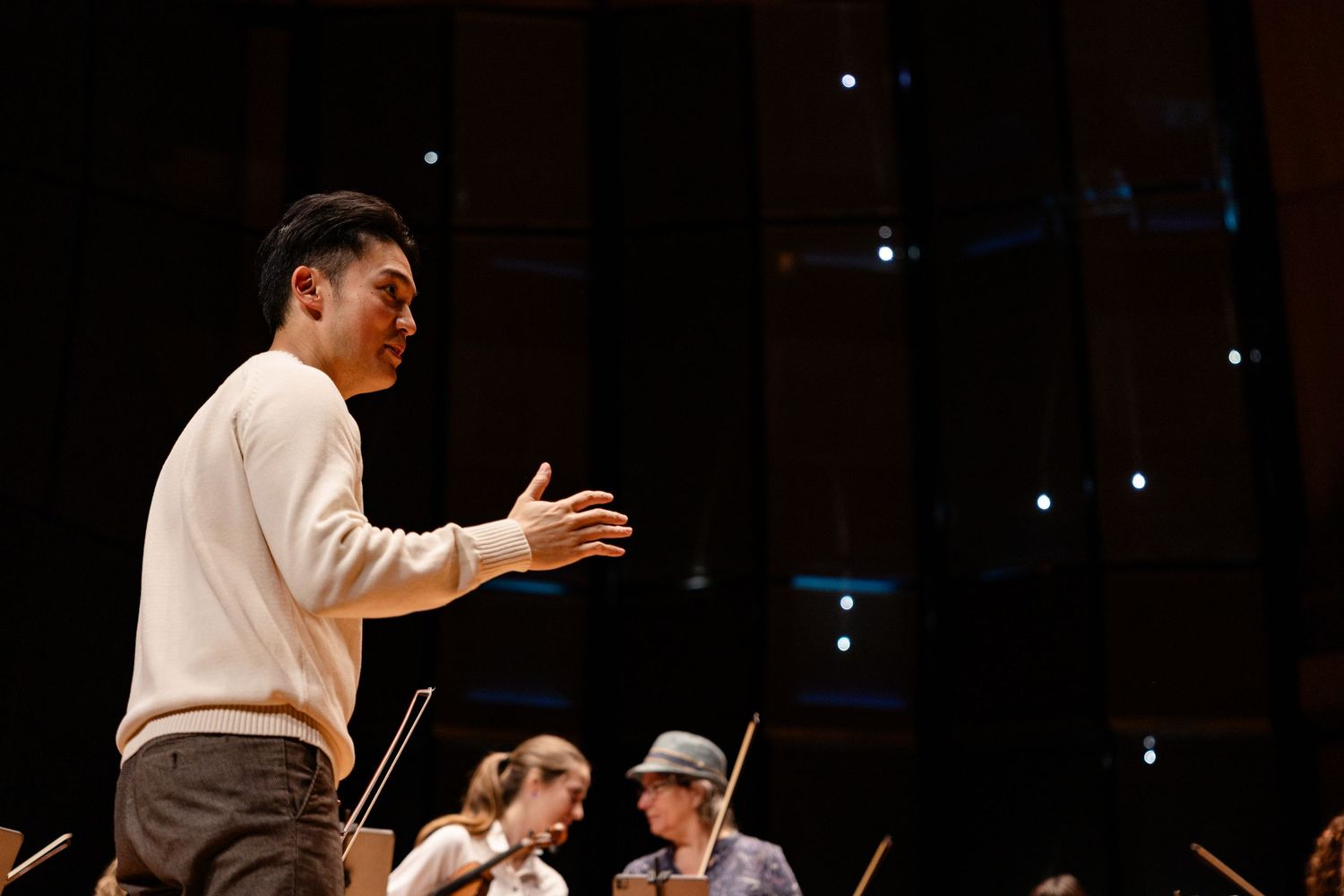 A conductor leads an orchestra rehearsal on a concert hall stage, gesturing expressively to musicians with string instruments.