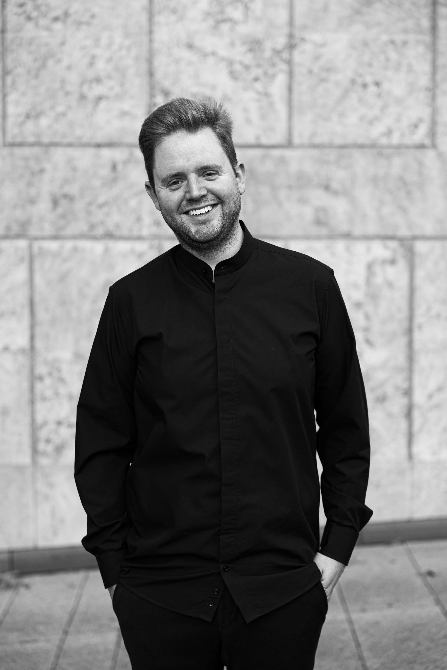Smiling man in black shirt standing confidently against a marble wall in a black and white portrait photo.