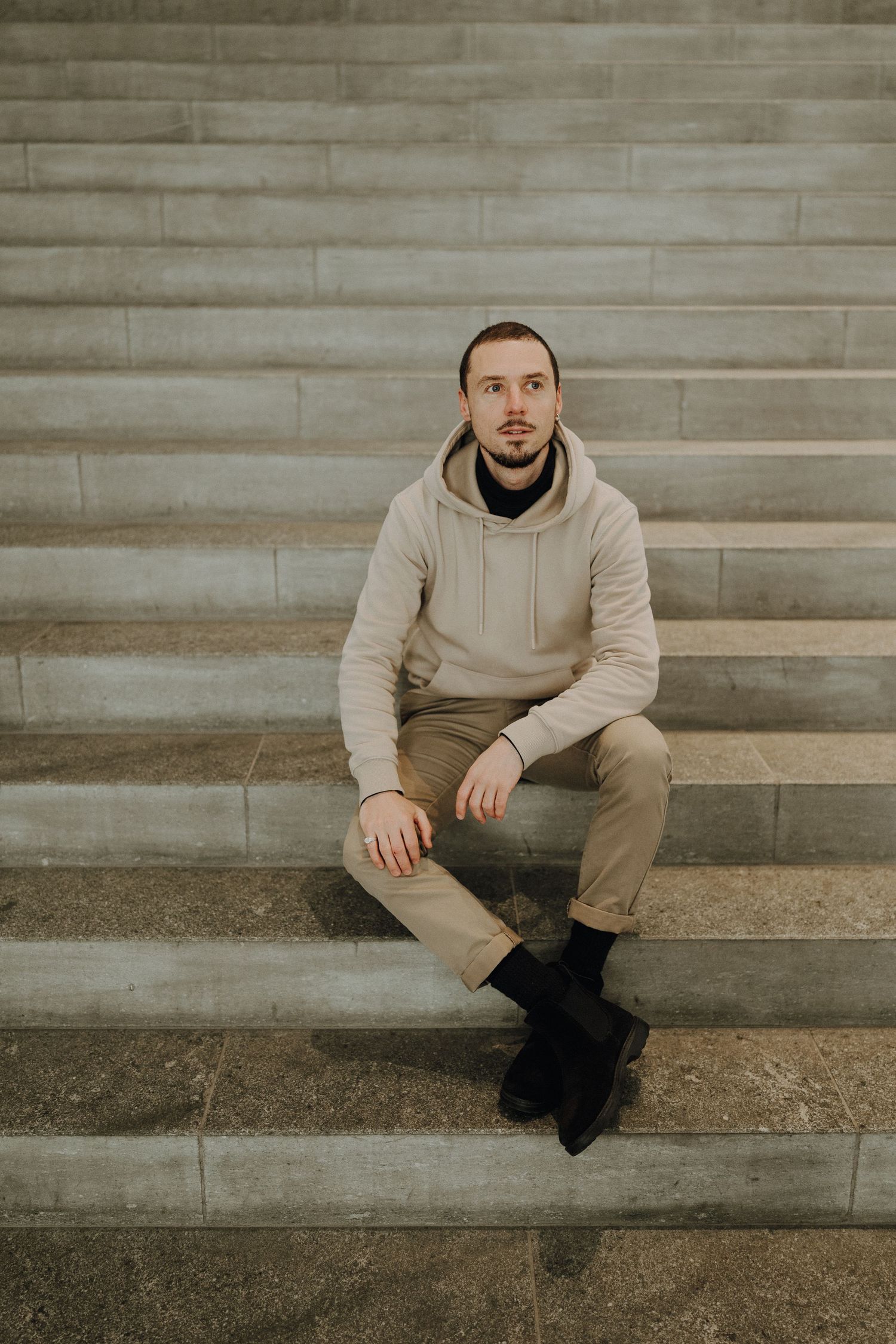 Young man in beige hoodie and khaki pants sitting on stone steps, looking at camera.
