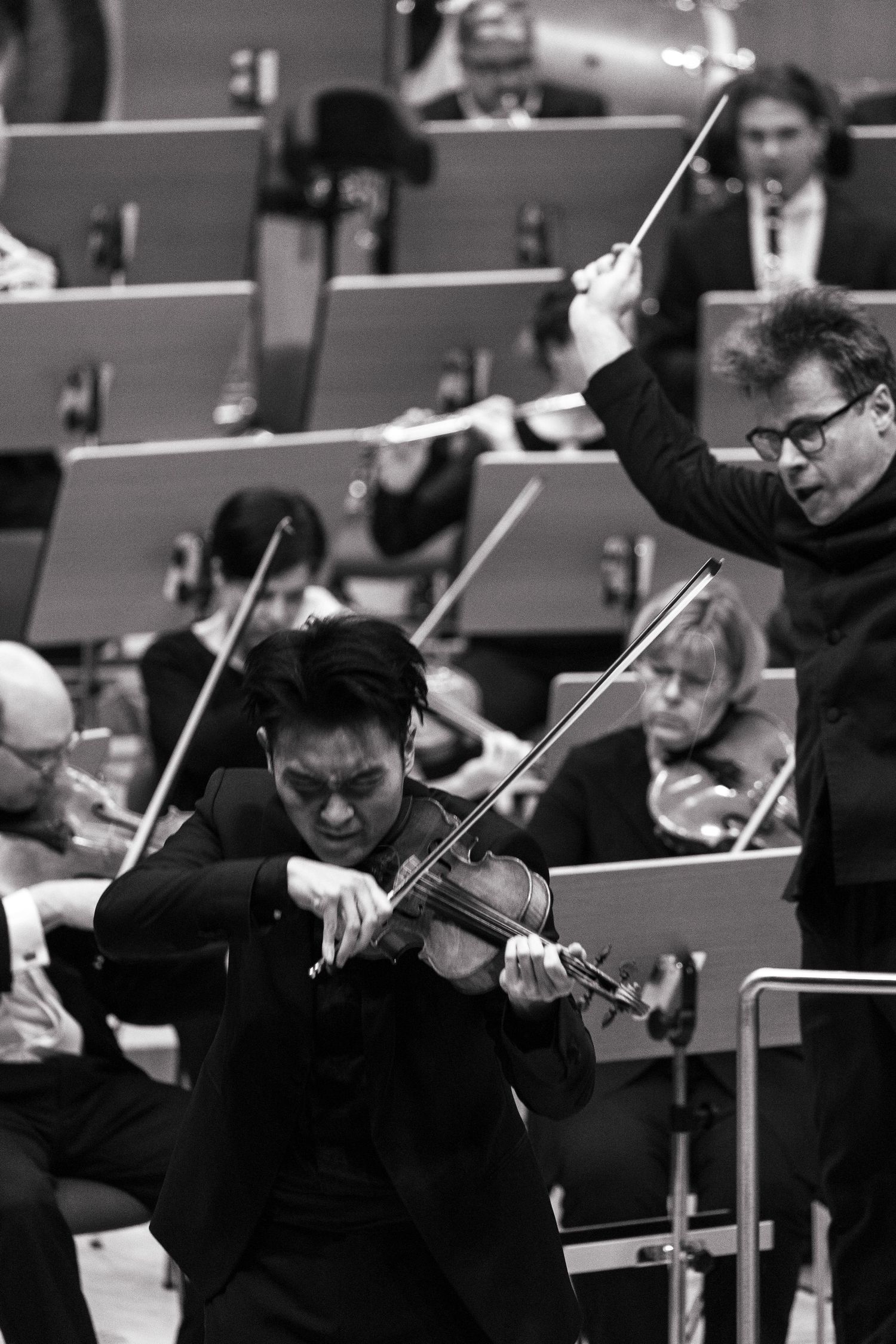 Black and white photo of a violinist performing with an orchestra conductor raising baton in background.