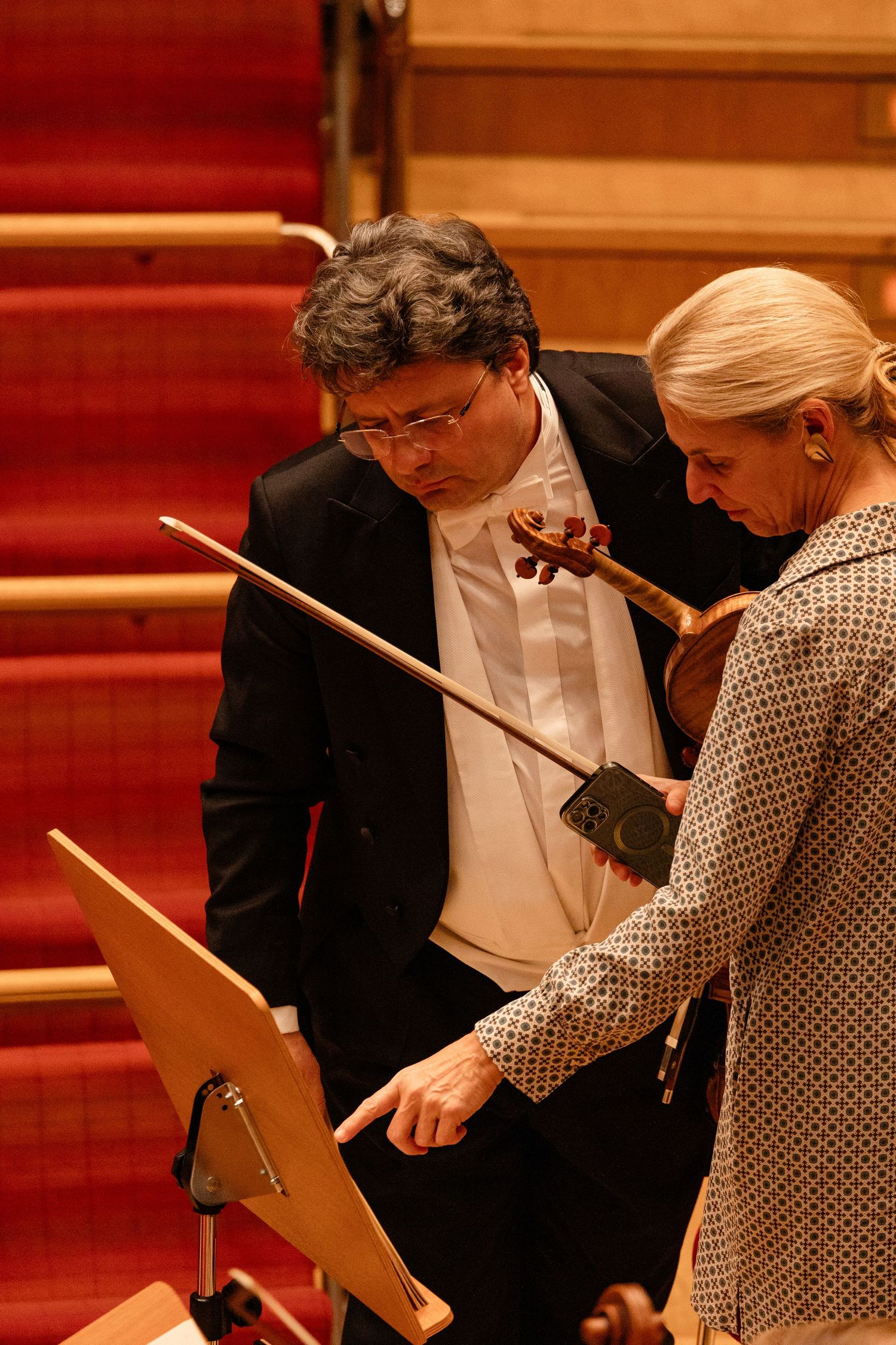 Musician in formal attire holds violin while reviewing sheet music on stand with colleague in concert hall.