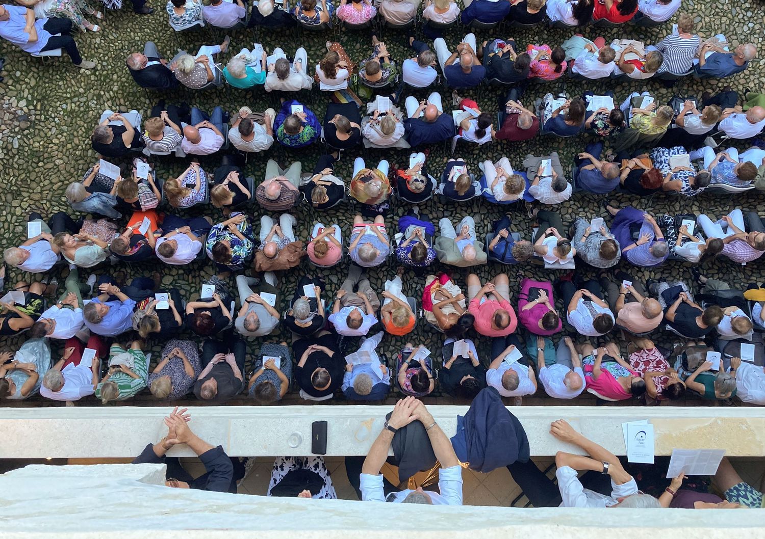Aerial view of a large crowd of people sitting in bleachers, many holding phones, at an outdoor event.