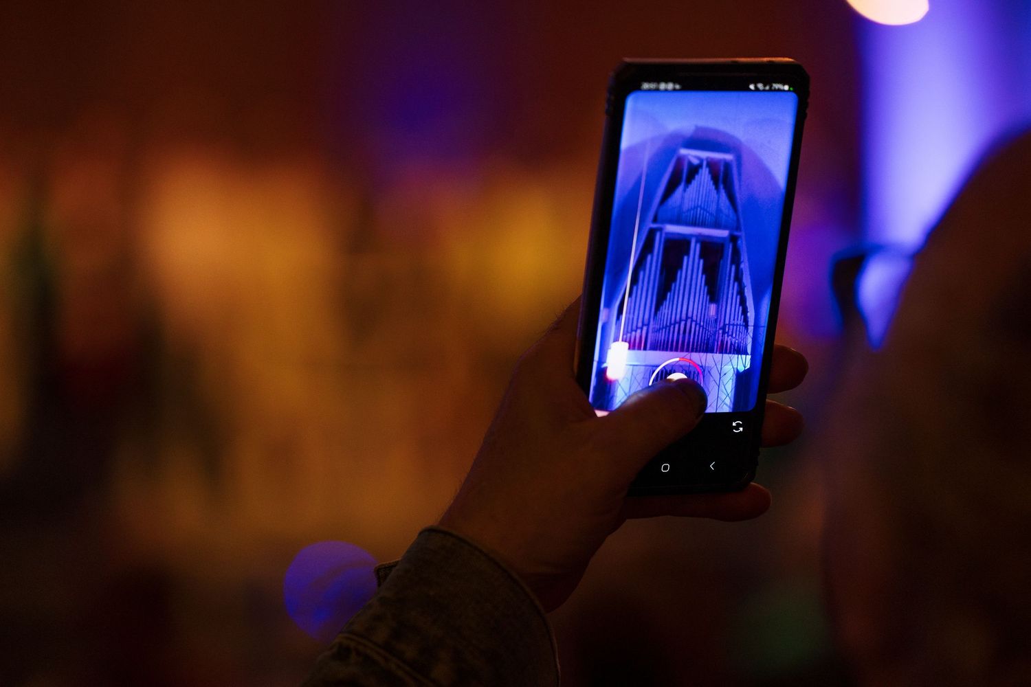 Person holding smartphone photographing illuminated display at dark event with colorful purple and blue lighting.