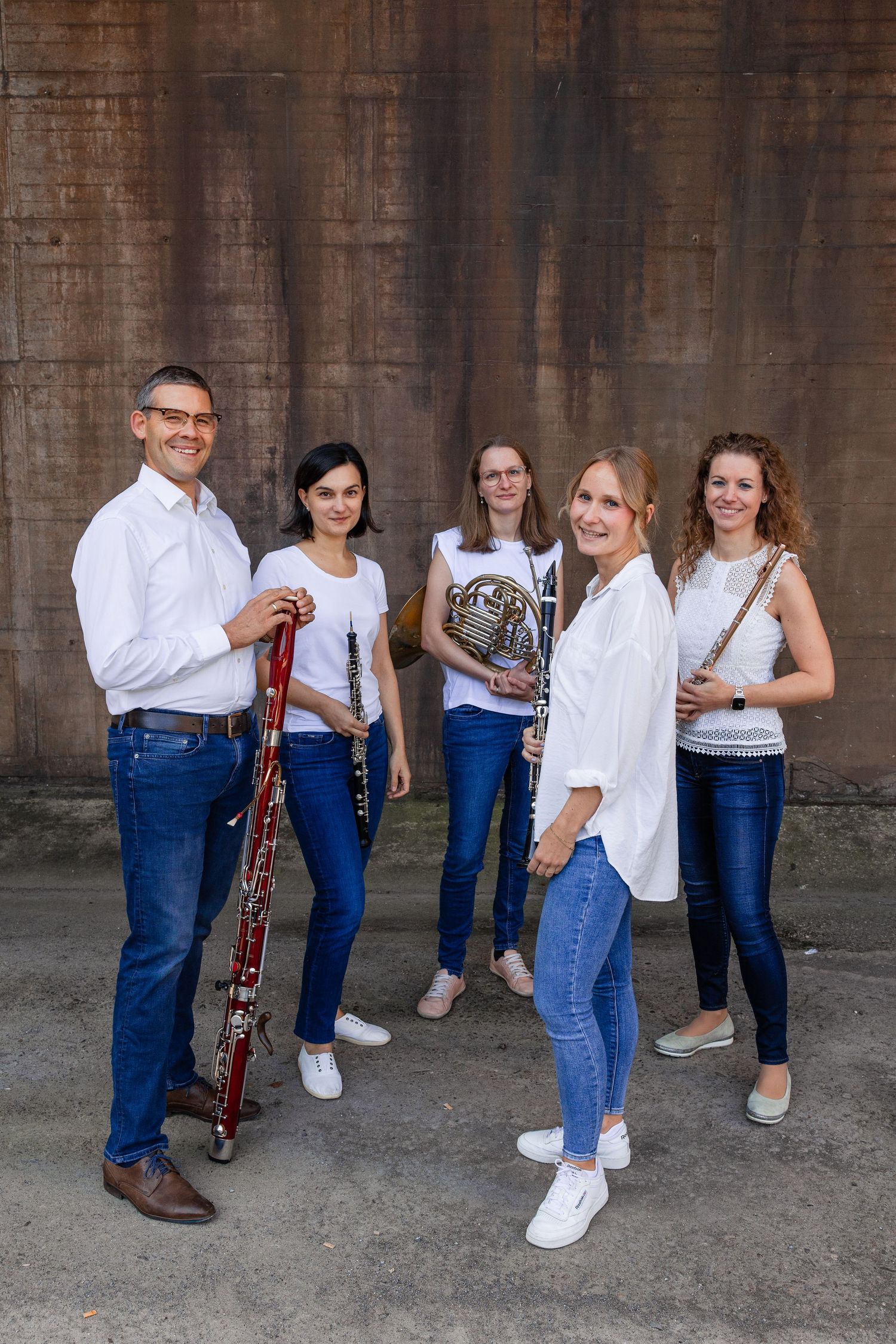 Five musicians in white shirts pose outdoors holding woodwind instruments including bassoon, clarinet, and French horn.
