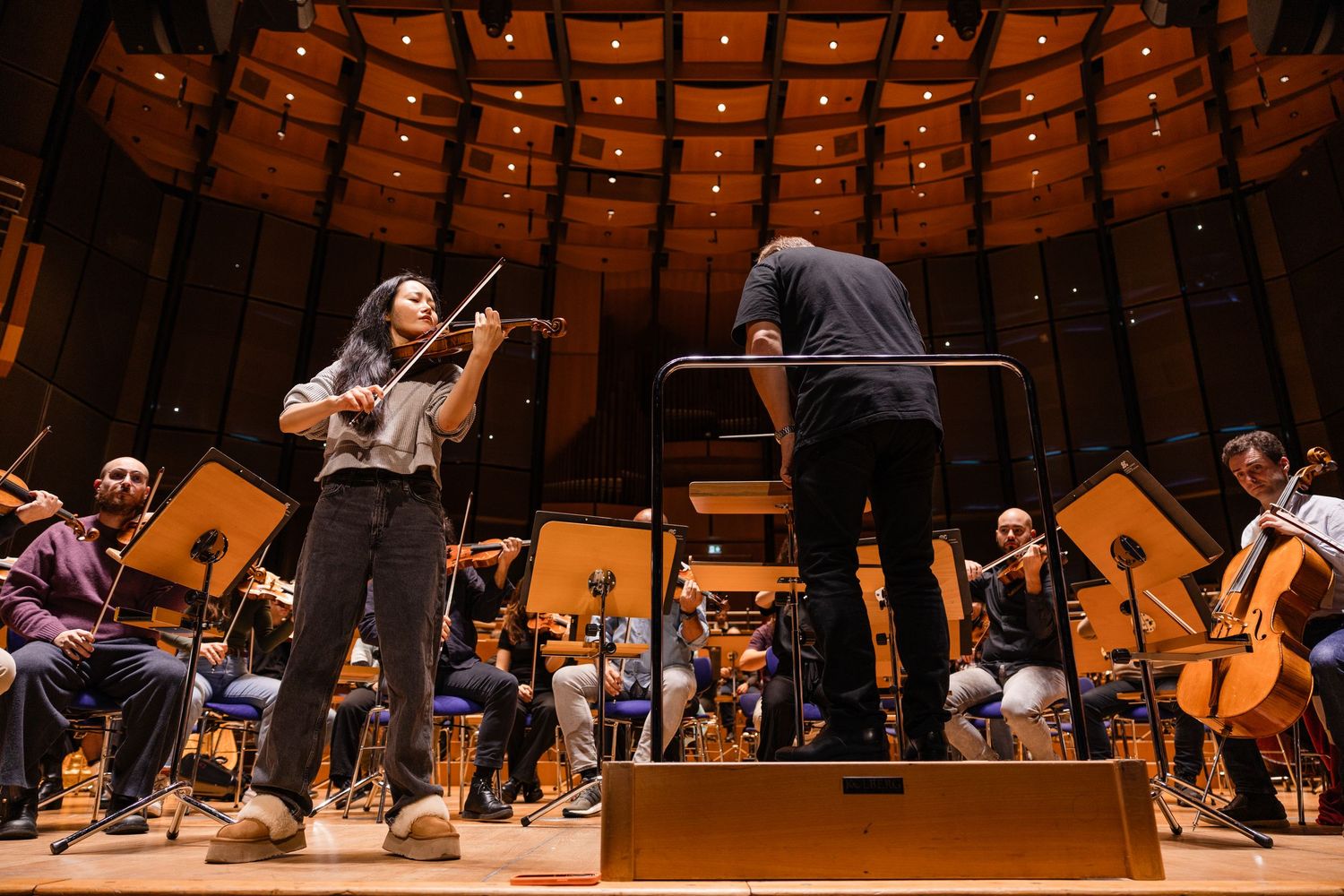 Violinist performs with orchestra conductor during rehearsal in a modern concert hall with warm wood paneling.