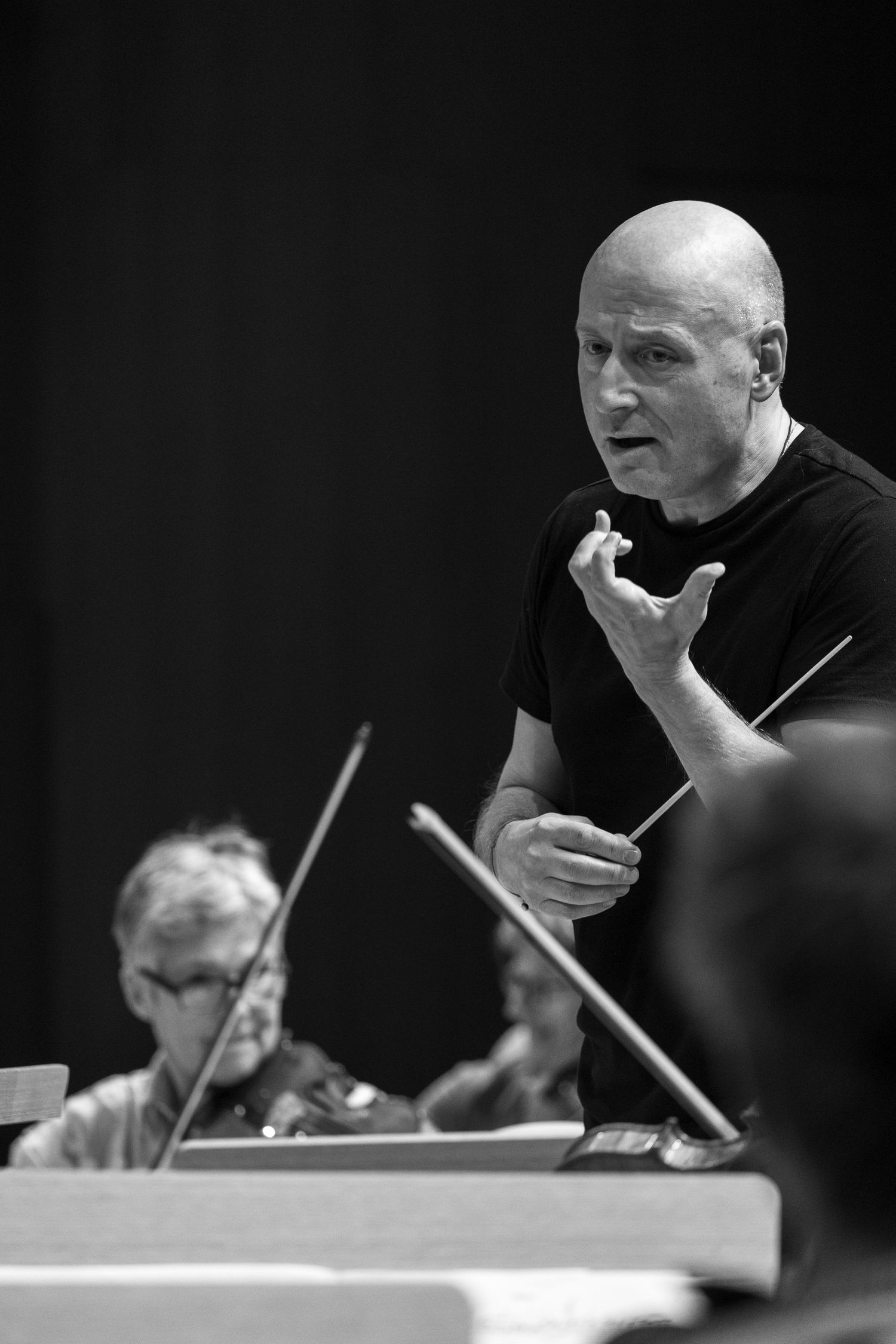 Bald conductor holding a baton gestures expressively during an orchestra rehearsal in black and white.