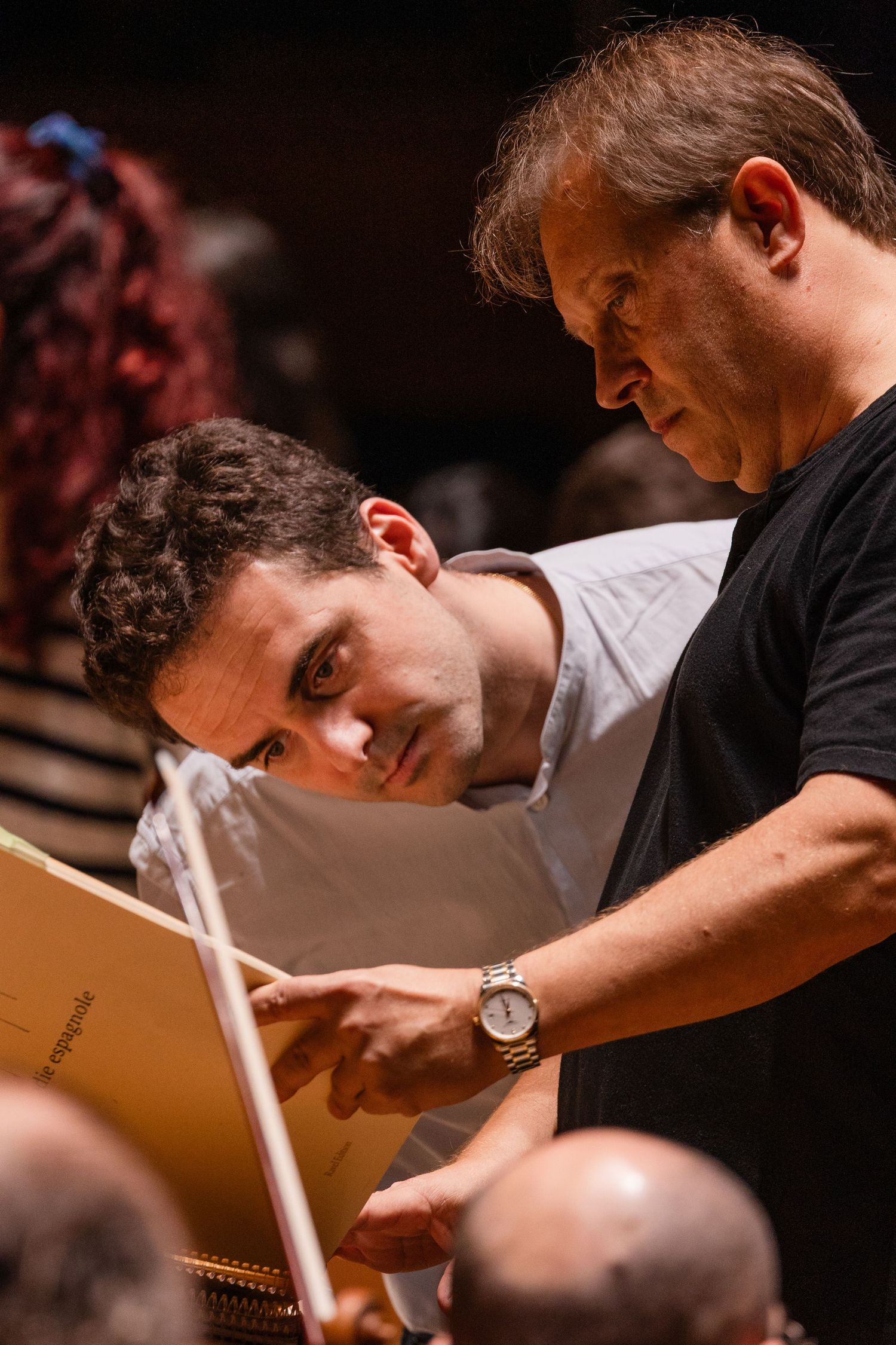 Two musicians review sheet music together during an orchestra rehearsal, one holding a conductor's baton.