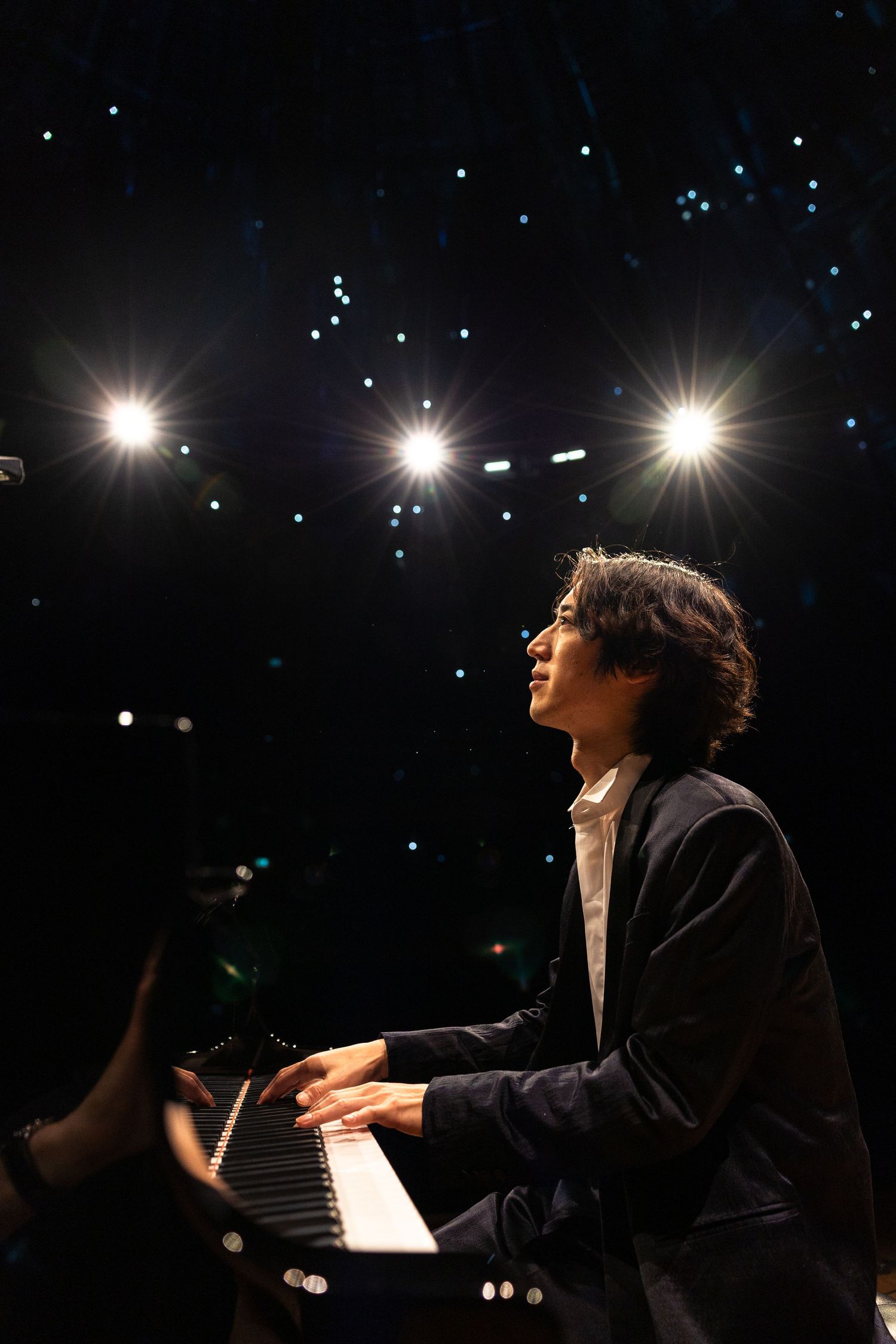 Pianist performing on stage under bright spotlights in a dark concert hall, playing grand piano keys with focused expression.