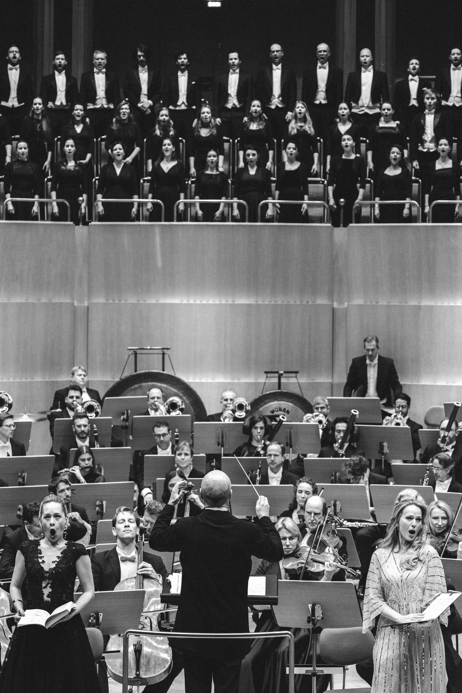Black and white photo of a full orchestra and choir performing on stage with a conductor and soloists in formal attire.