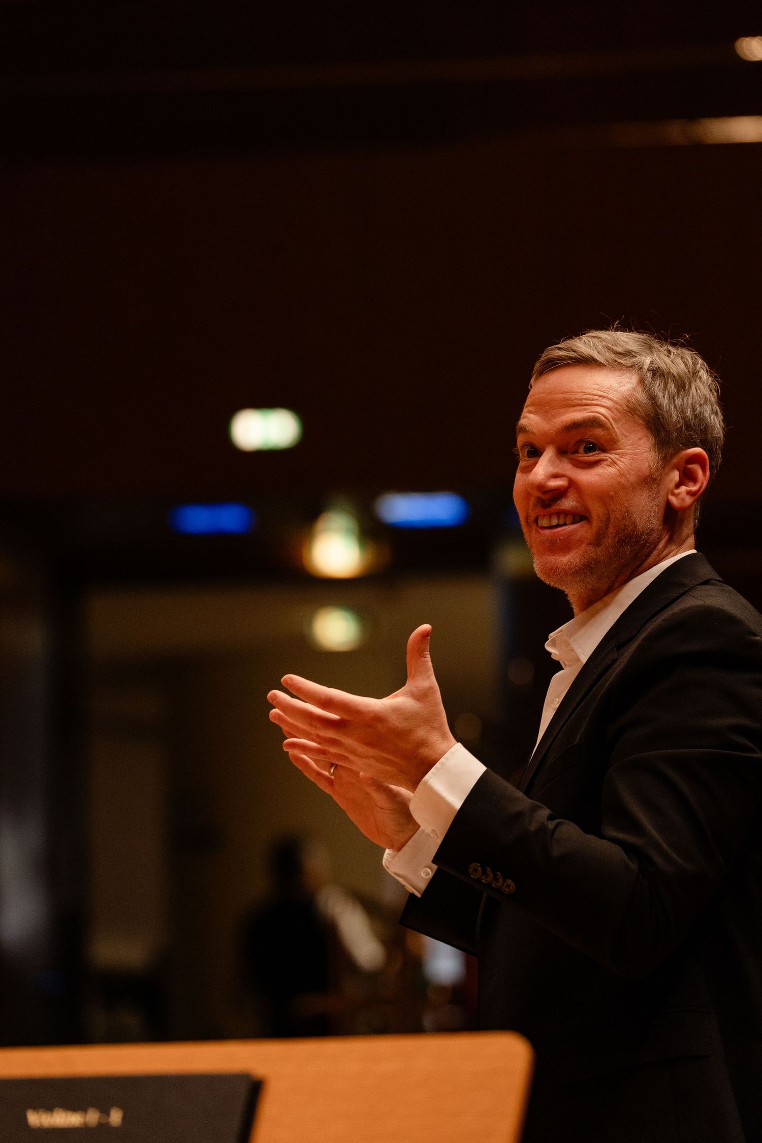 A smiling man in a black suit conducts an orchestra, gesturing expressively with his hands in a concert hall.