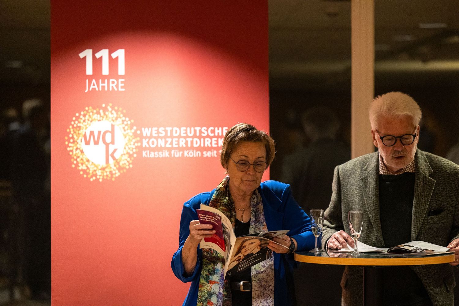 Two attendees browse programs at a Westdeutsche Konzertdirektion 111th anniversary event in an elegant venue.
