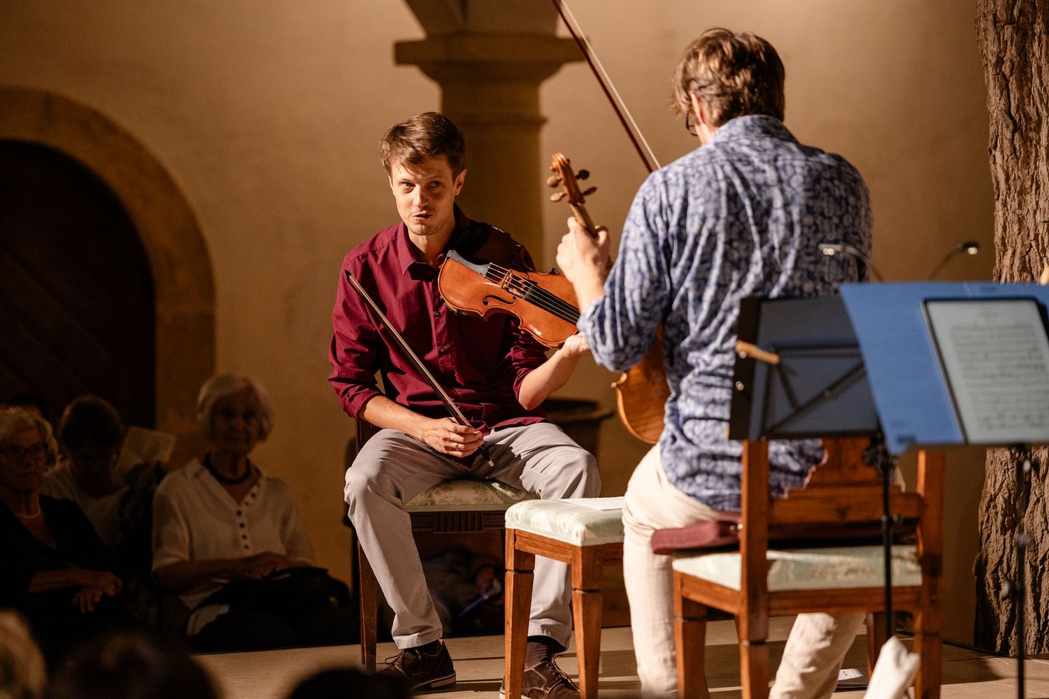 Two musicians performing violin in a warmly lit concert hall with an audience seated in the background.