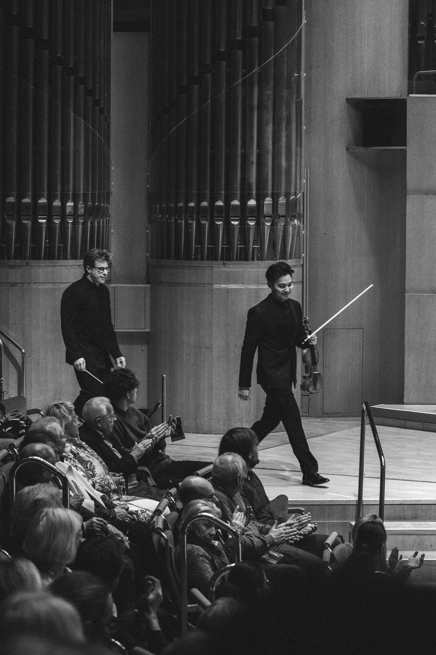 Two musicians in black suits walk on stage holding violin bows at a classical concert hall performance.