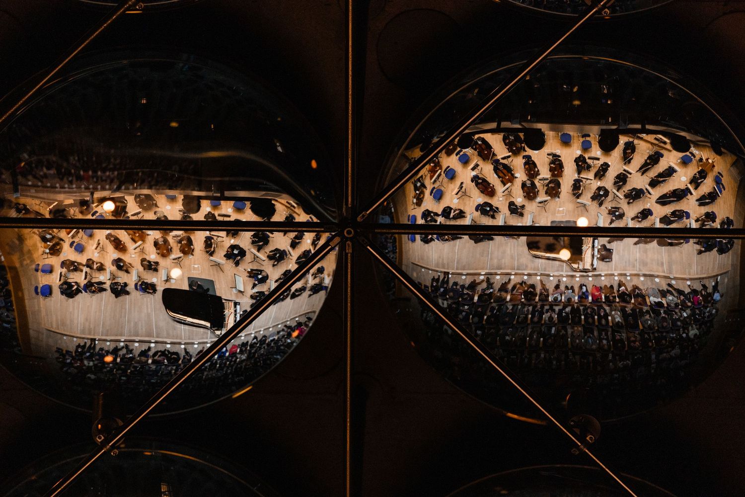 Aerial view of a packed concert hall audience reflected in two circular mirrors creating a dramatic split composition.