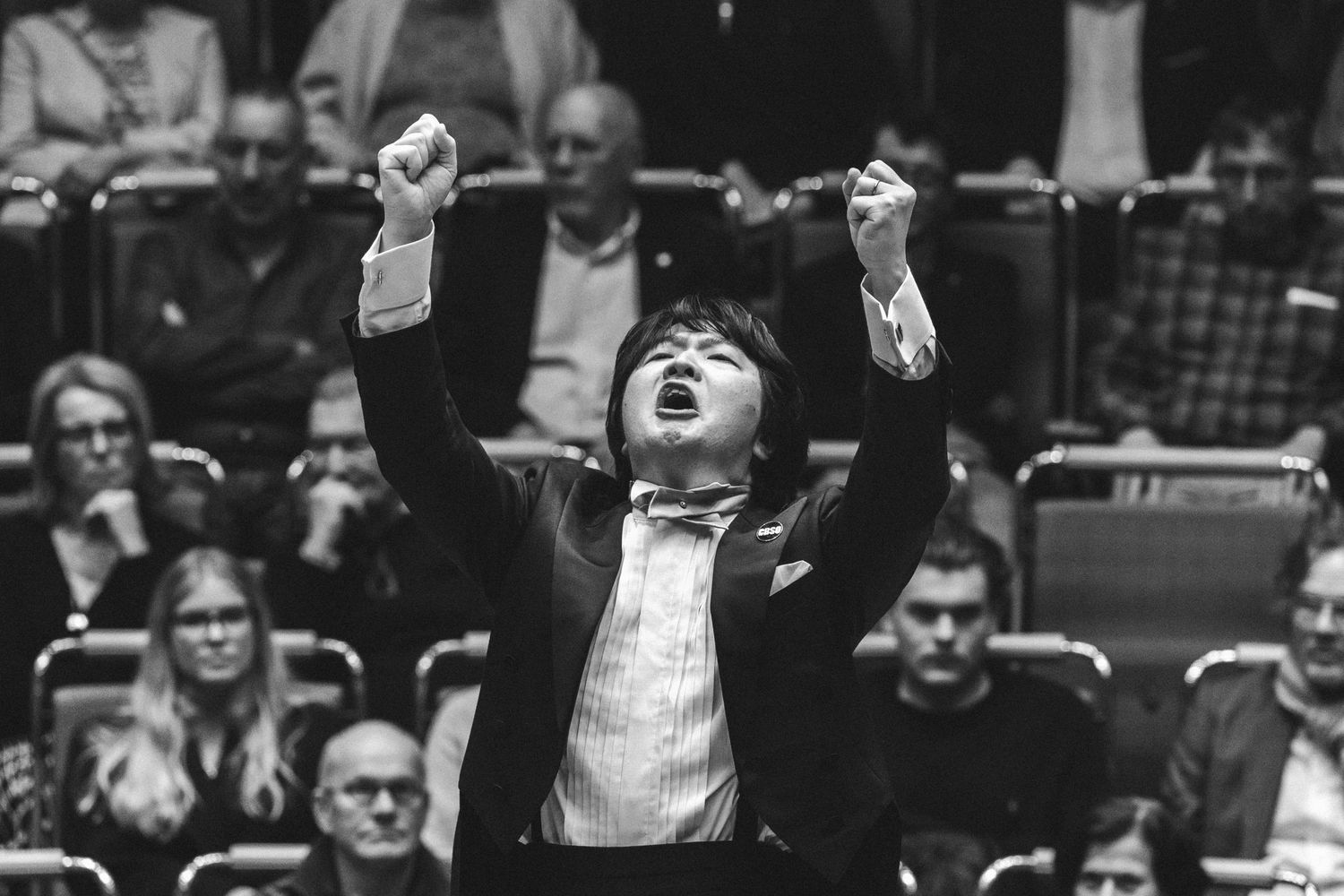 Conductor passionately raises fists during orchestral performance in black and white concert hall photo.