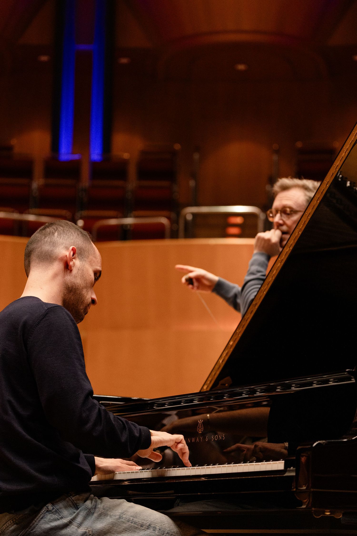 Pianist playing grand piano during rehearsal while conductor points directions in concert hall.