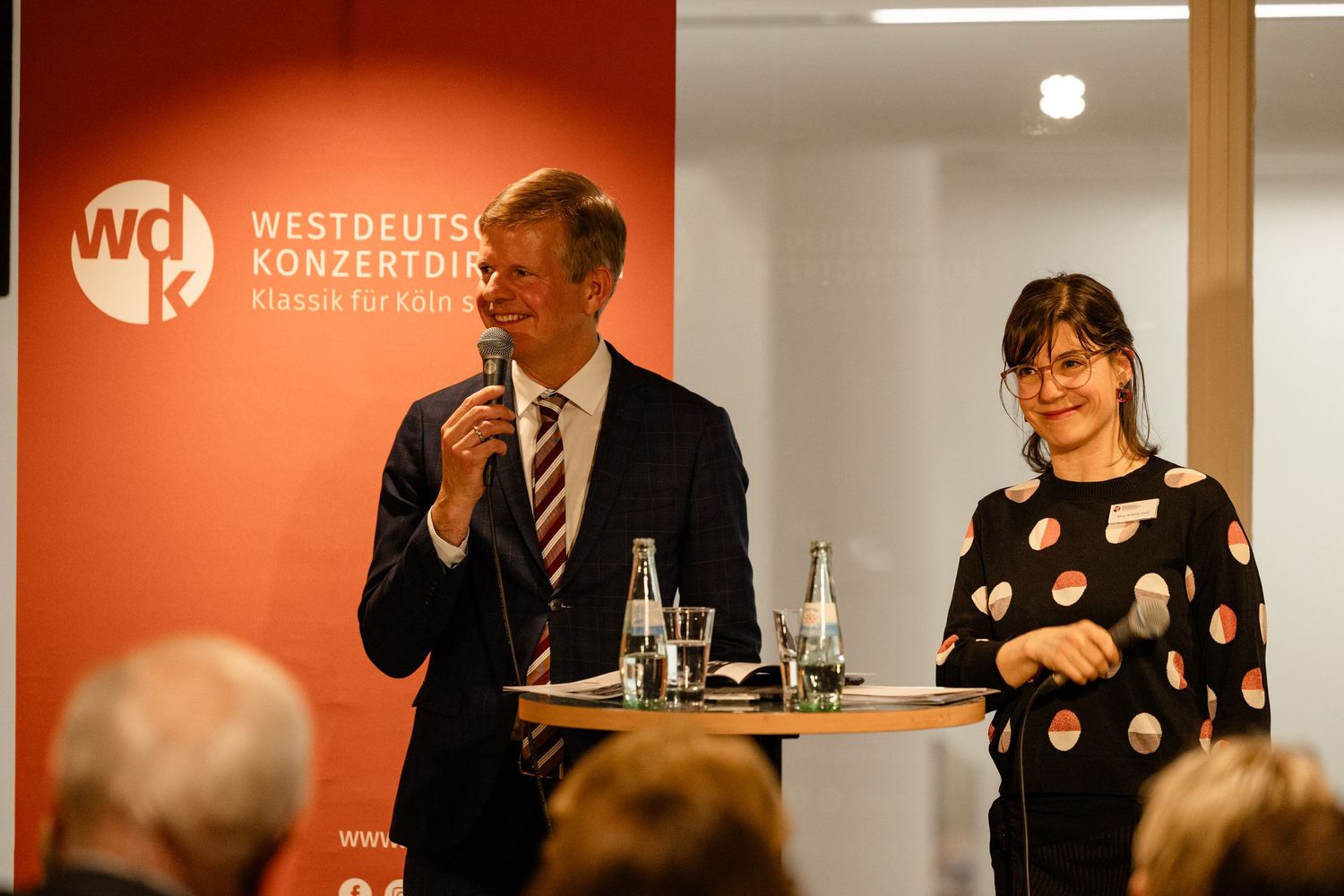 Two speakers present at a Westdeutsche Konzertdirektion event, man in suit holding microphone beside woman in polka dot top.