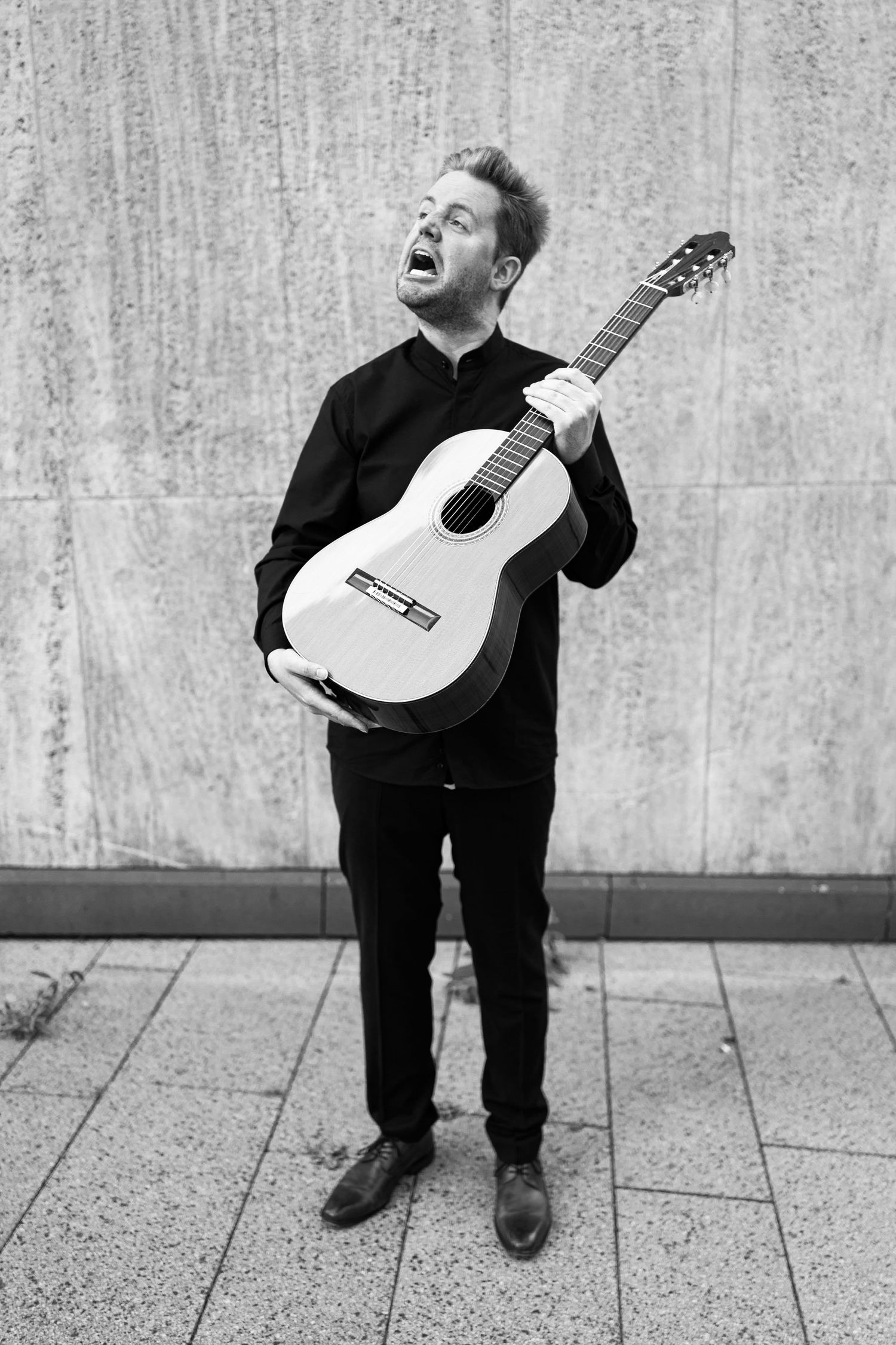 Man in black outfit passionately playing acoustic guitar outdoors, black and white photo.
