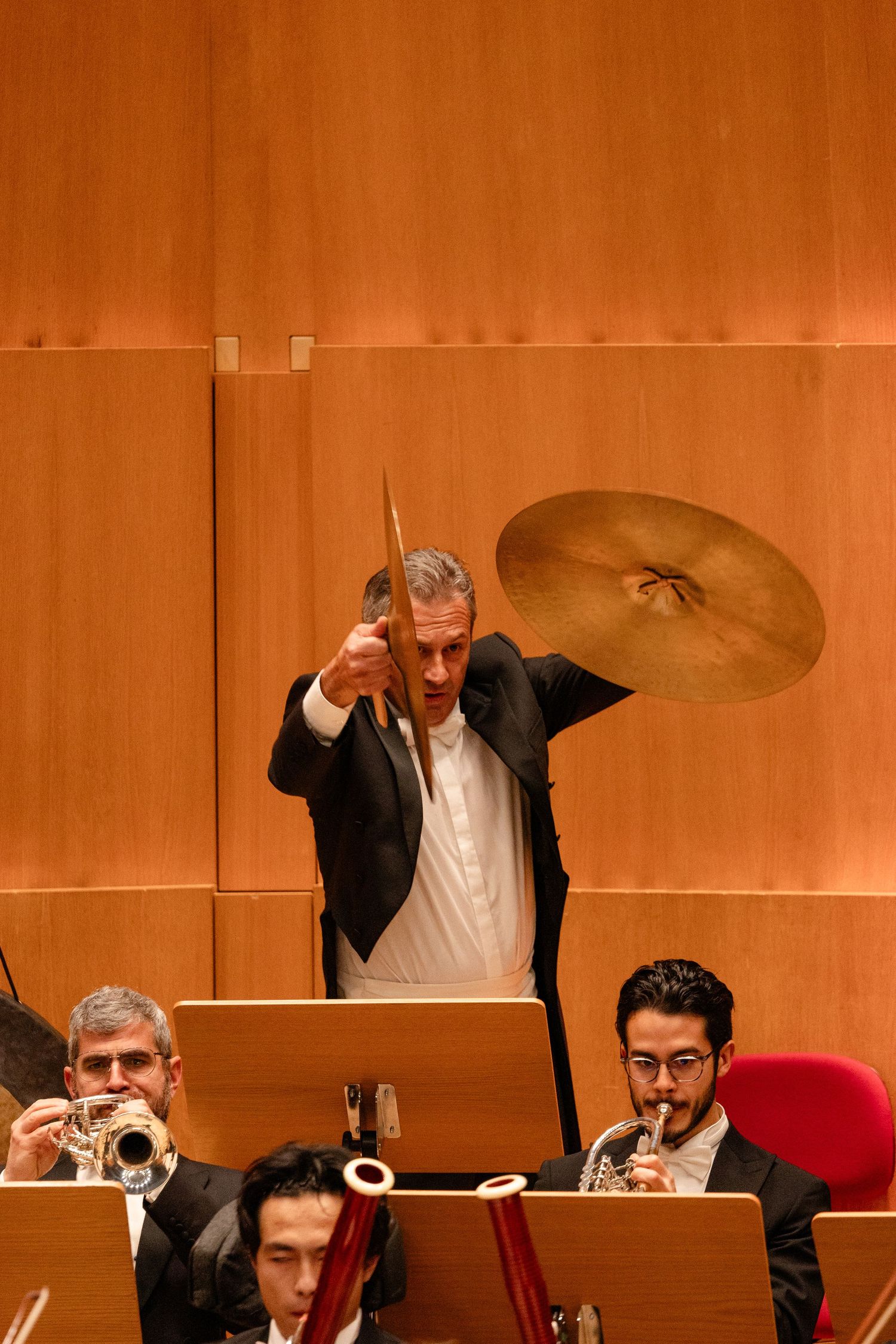 Musicians in formal attire perform on stage with brass instruments and cymbals during an orchestral concert.