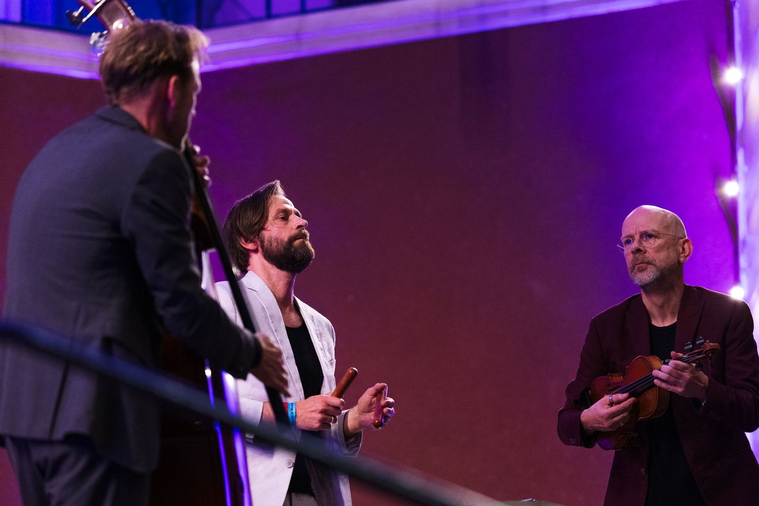 Three men engaged in conversation on a purple-lit stage, one holding a guitar, dressed in smart casual attire.