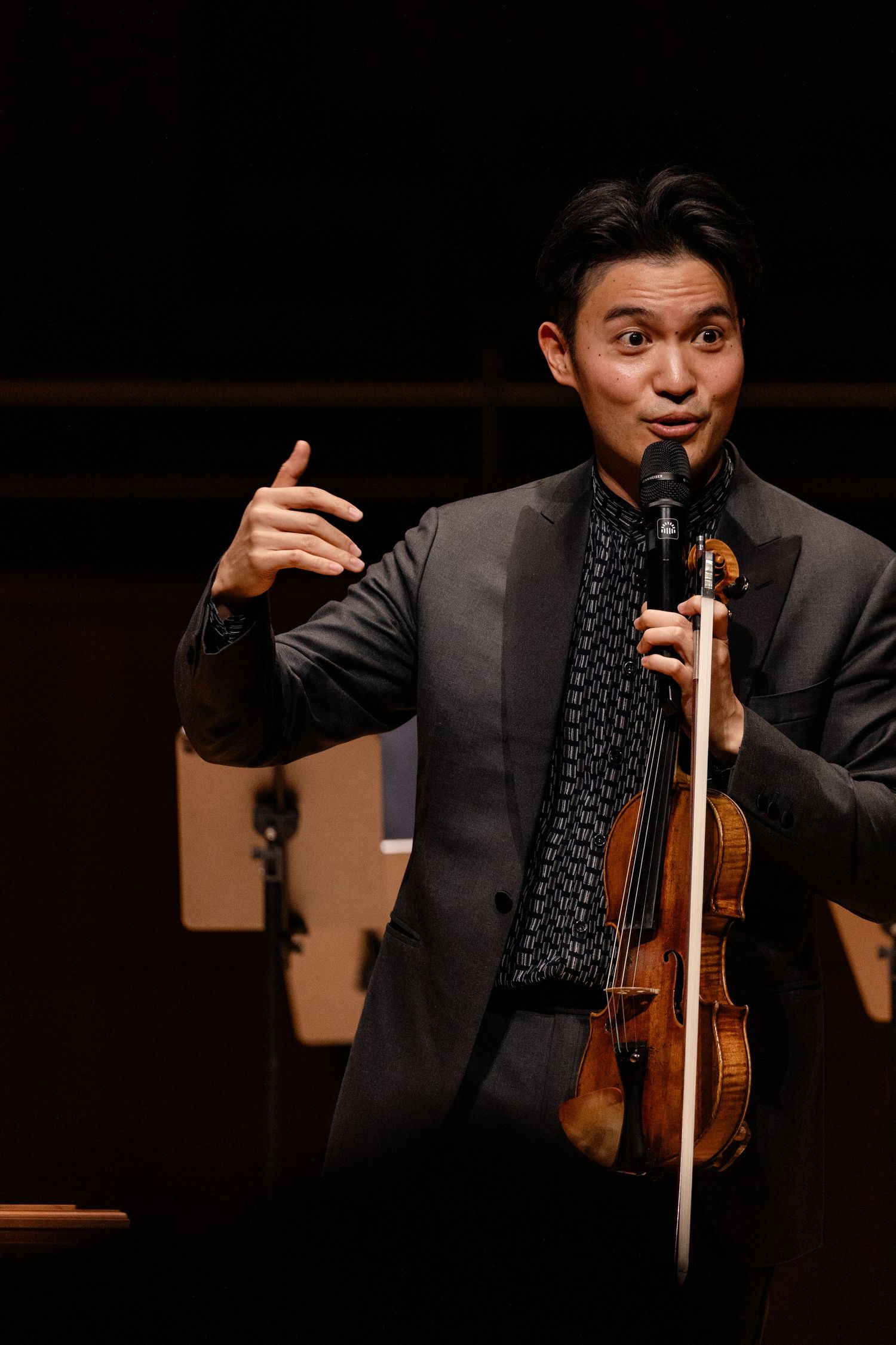 Violinist in dark suit speaks at microphone on stage, holding violin and bow during a live performance.