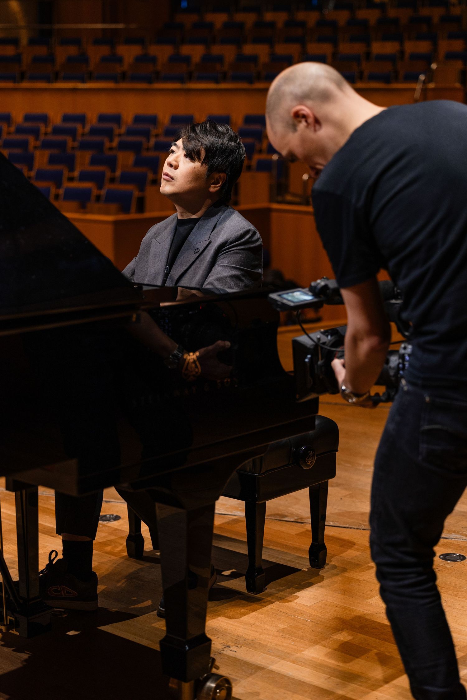 Pianist performing on grand piano on concert hall stage while videographer films the session.