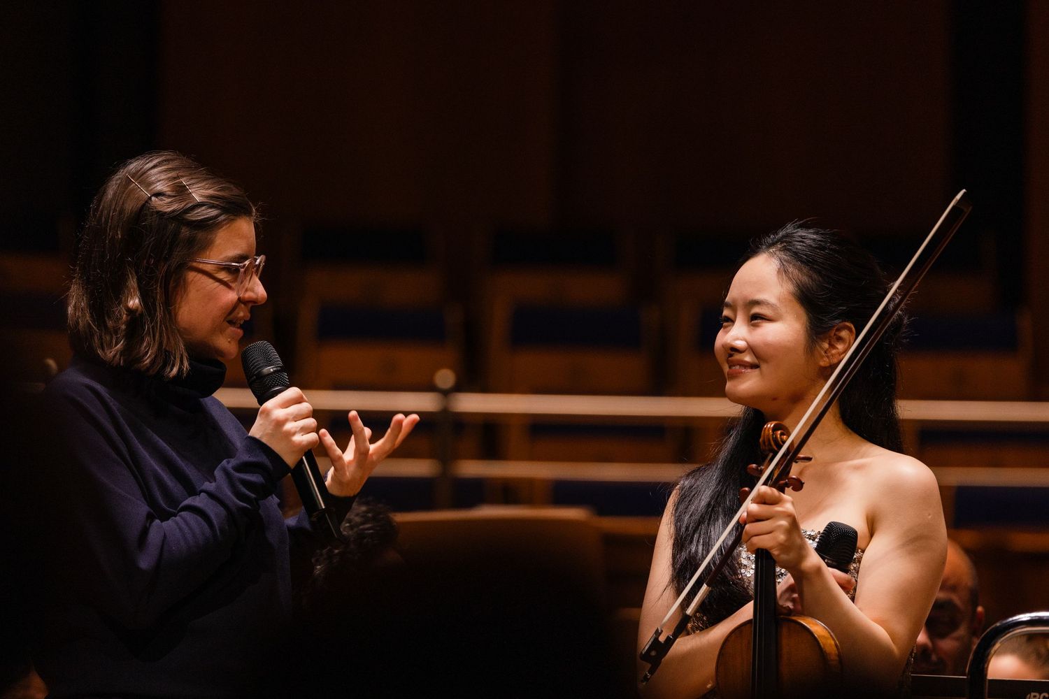 Two musicians converse on stage, one holding a violin and bow, the other speaking into a microphone in a concert hall.