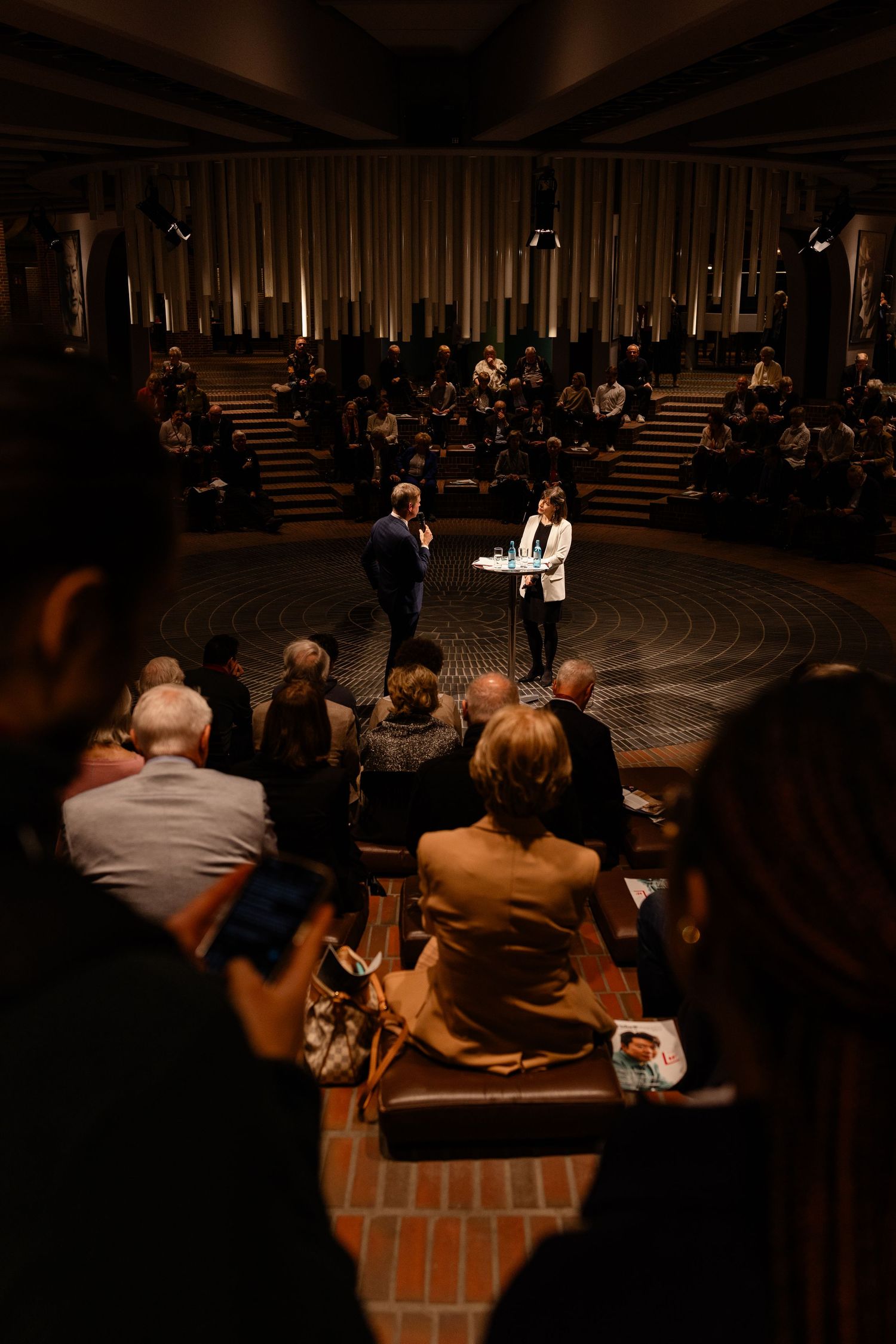 Two speakers present to an engaged audience in a dimly lit circular venue with elegant columns and tiered seating.