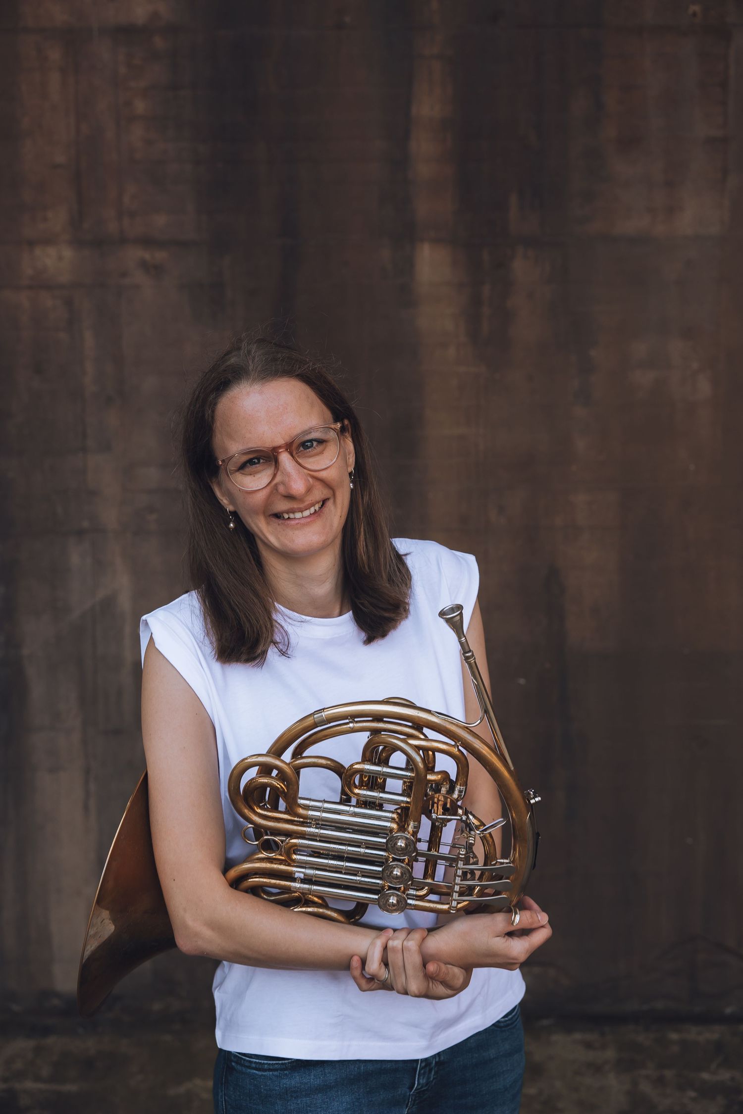 Smiling woman with glasses holding a French horn, wearing a white sleeveless top, standing against a rustic brown wall.
