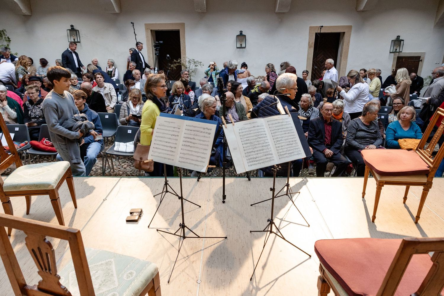 Music stands with sheet music face a seated audience in an elegant hall during a classical chamber music concert.