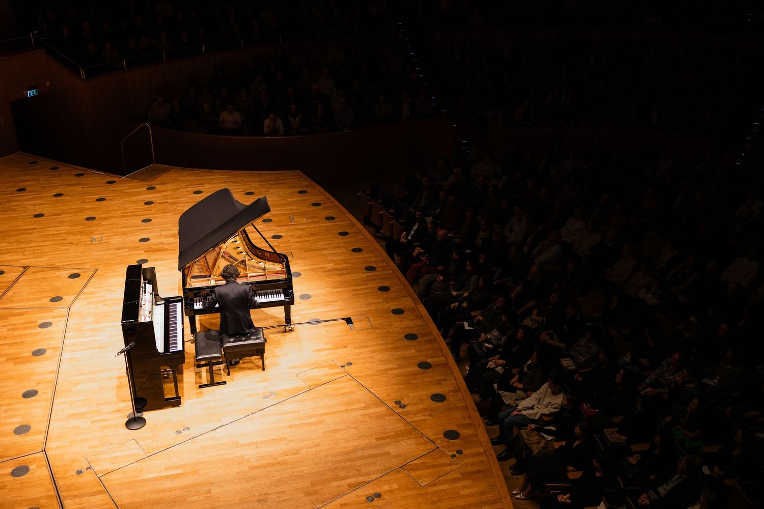 A grand piano spotlit on a wooden concert hall stage, viewed from above with audience seated in the background.