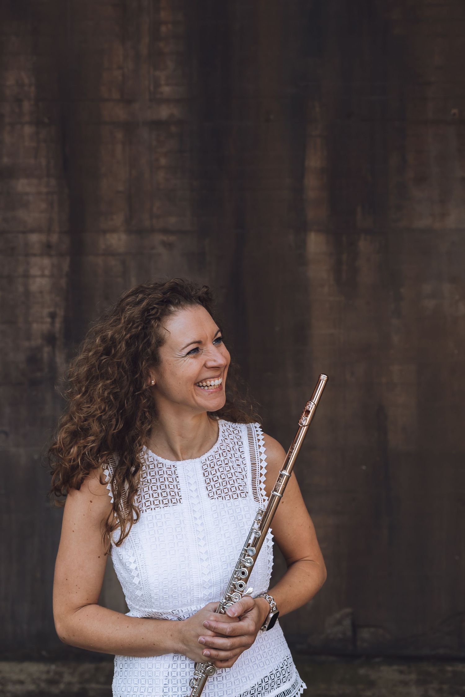 A smiling woman with curly hair holds a silver flute, wearing a white lace dress against a rustic brown wall.