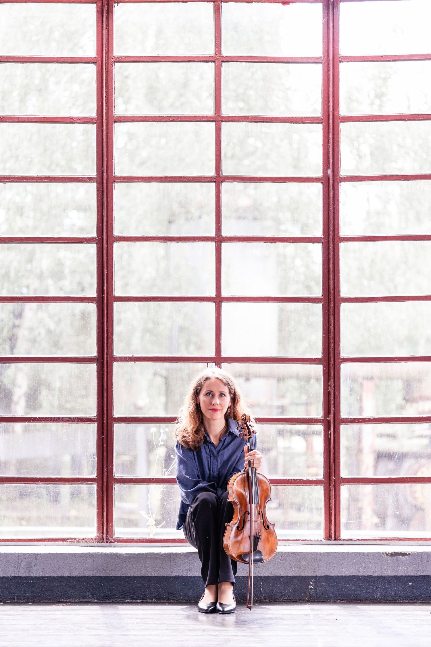 A woman with curly hair sits holding a violin and bow in front of a large red-framed window in a bright room.